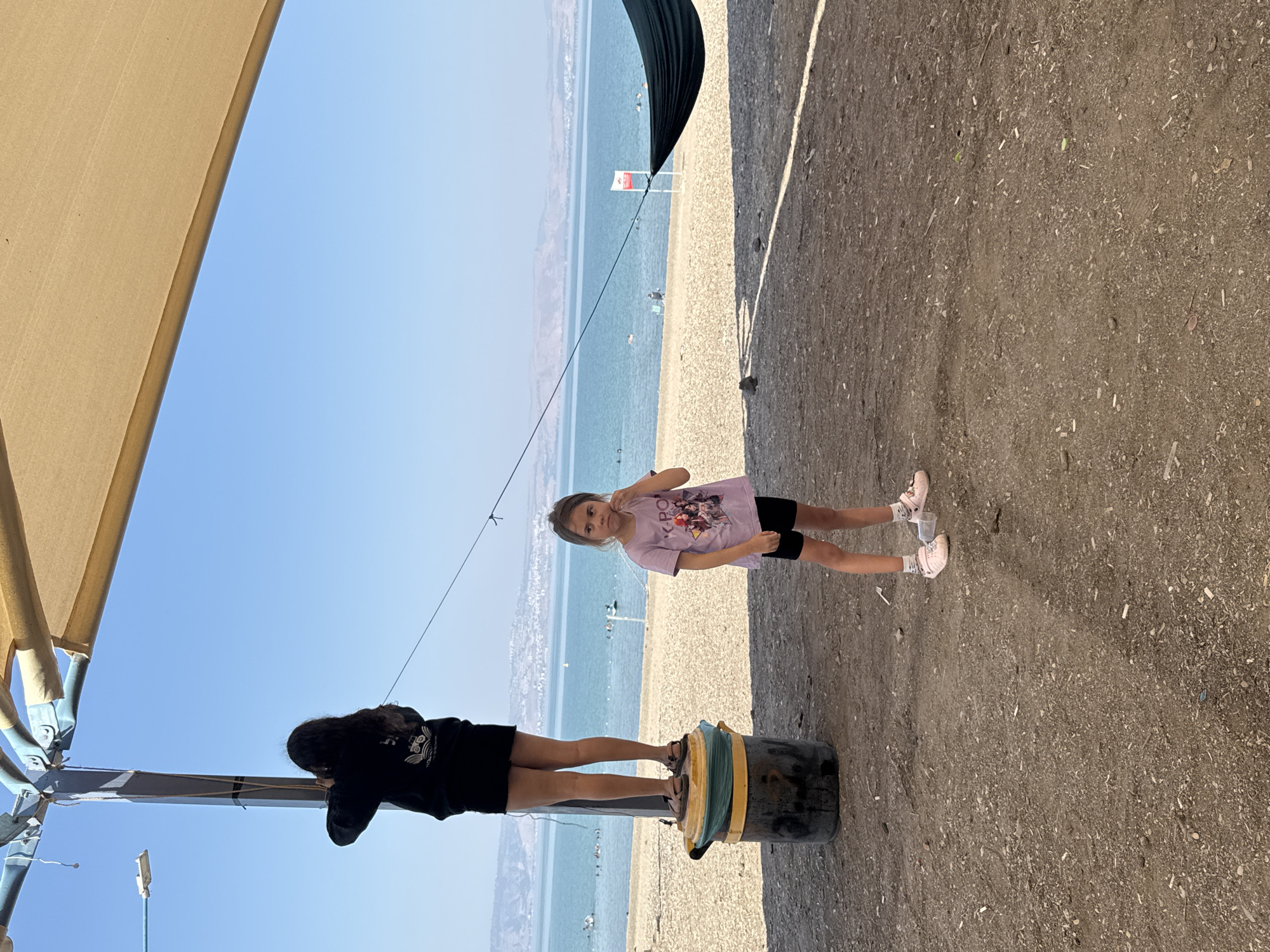 This image depicts a scene at a beach on the Sea of Galilee, located in the Golan Regional Council, Israel. Two young girls are the central subjects. The girl on the left, with long dark hair, stands on a large cylindrical object, possibly a repurposed bin, wearing a black hooded sweatshirt and a black skirt. She is looking towards the left and upwards, her hand near her face. To the right, a younger girl stands on the sandy ground, facing forward, wearing a purple t-shirt with graphics and black shorts. She has her hand on her chin, appearing thoughtful or pensive.
The background reveals the Sea of Galilee, with calm blue water extending to a distant shoreline lined with hills and a town or city. Several people are visible in the water, swimming or engaging in recreational activities. A black hammock is suspended between two points, partially visible on the right side of the image. The foreground is a mix of gravel and dirt, with some debris scattered on the ground. Above, a beige canopy stretches across the top of the frame, supported by a metal structure. The sky is clear and blue. The overall impression is a casual, outdoor setting by a body of water on a sunny day.