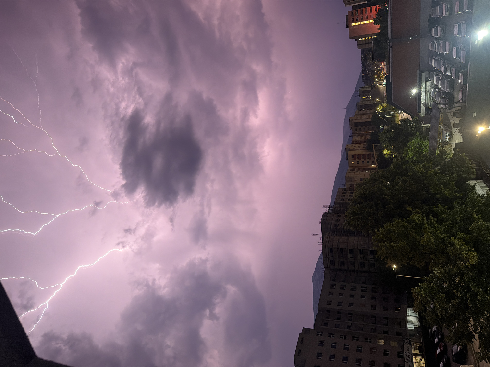The image captures a dramatic thunderstorm over Sofia, Bulgaria, likely during twilight. Jagged bolts of white lightning illuminate a heavy, purple-pink sky filled with dark, ominous clouds. Below, the urban landscape of Sofia is visible, characterized by rows of apartment buildings of varying heights and architectural styles. Some buildings appear modern, while others show signs of older construction, with some undergoing renovation. Trees with dark green foliage dot the scene, interspersed with streetlights casting a yellowish glow. A parking lot filled with numerous cars is visible in the foreground, along with a covered parking area. In the distance, rolling hills or mountains form a silhouetted backdrop against the tempestuous sky. The overall impression is one of a powerful natural event unfolding above a dense urban environment.