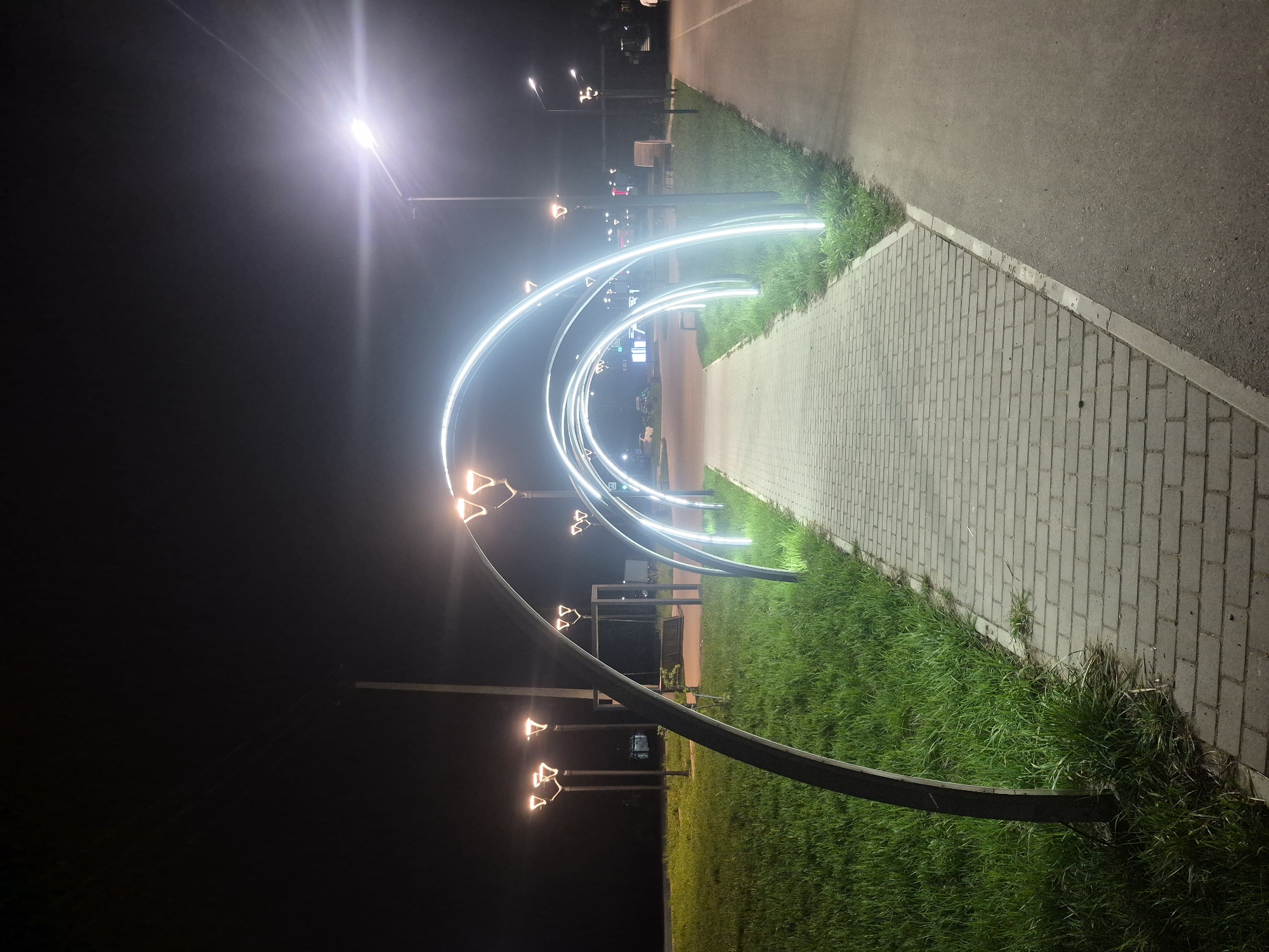 At night in Chaikovsky, Russia, a pedestrian walkway is illuminated by an archway of glowing white lights. The walkway is composed of gray interlocking pavers bordered by green grass on both sides. Adjacent to the paved walkway is an asphalt path. Dark metal arches support the glowing lights, and streetlights with orange lighting fixtures are visible in the background. The environment is dark, suggesting a nighttime setting. The focus is on the architectural lighting and pedestrian infrastructure.