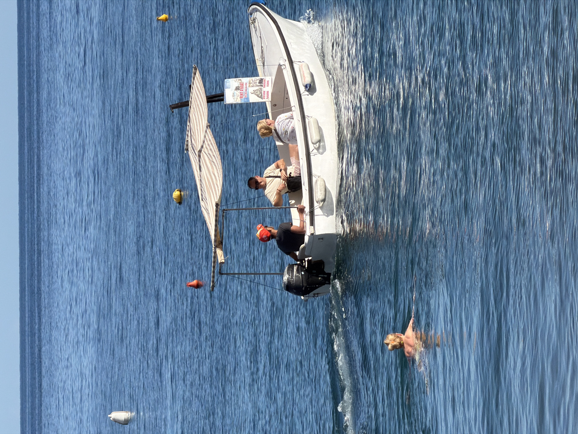 The image captures a marine scene on a sunny day. A small white boat with three people aboard is moving on the water, leaving a foamy wake. One person is operating the motor, another is standing nearby, and a third is positioned in the front of the boat. A striped sail is attached to the boat. Floating in the water are several buoys, colored white, yellow, and orange. A woman is swimming in the water away from the boat. A sign with text is affixed to the boat. The scene is set in Petrovac centar, Montenegro.