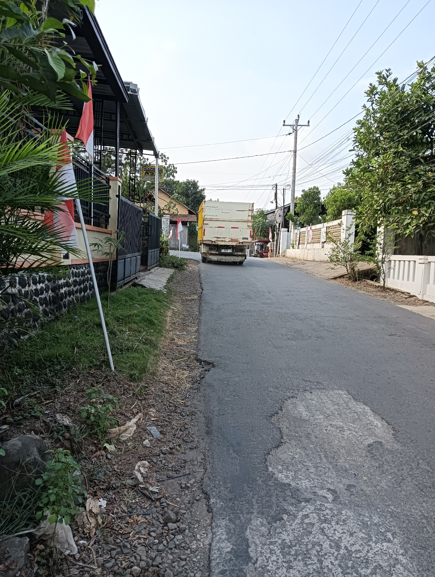 A truck drives down a road in Cluwak, Indonesia. The truck is traveling away from the viewer and is in the center of the frame. The road is asphalt and has some cracks and potholes. There are trees and houses on both sides of the road. There are also some Indonesian flags on the left side of the road. The sky is clear and blue.