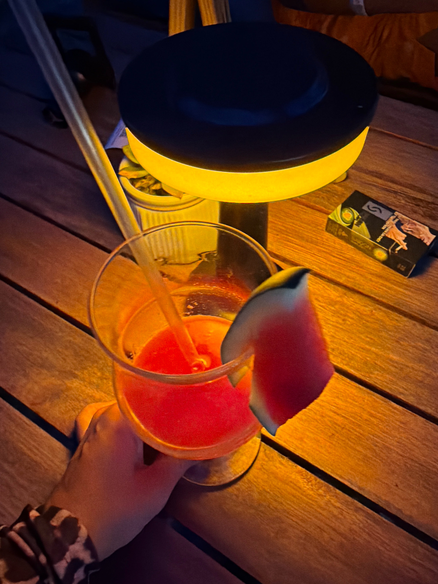 A hand with a patterned sleeve holds a clear glass of red liquid, garnished with a slice of watermelon. A clear straw extends into the glass. The glass rests on a wooden table, illuminated by a yellow-glowing lamp with a dark top. To the right of the glass, a pack of cigarettes with a design featuring hands and eyes is visible on the table. The scene appears to be outdoors at night, possibly in Nha Trang, Vietnam, given the warm ambiance and tropical elements suggested by the watermelon garnish.