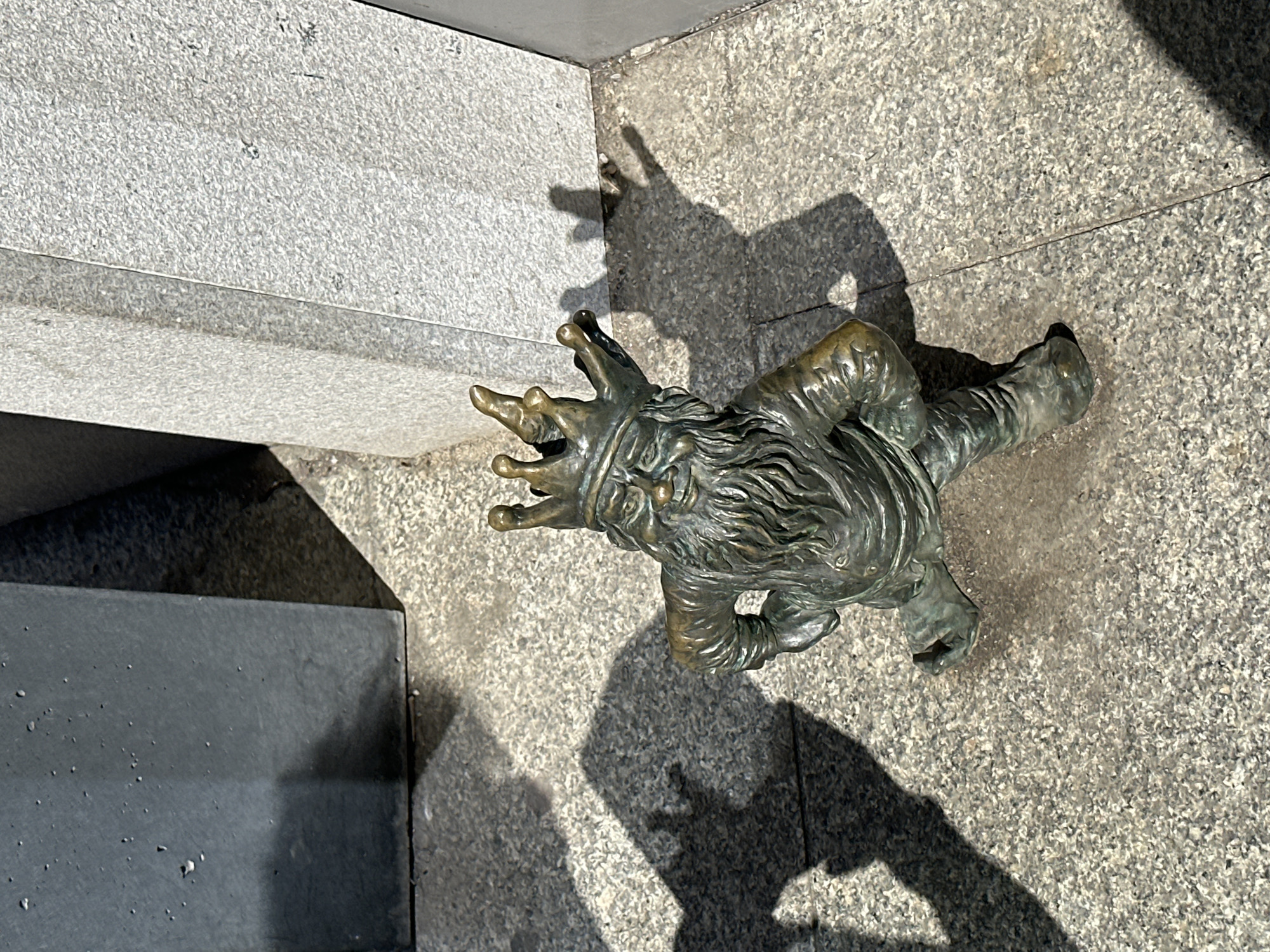 The image shows a bronze dwarf statue, a type of sculpture that is popular in the city of Wrocław, Poland. The dwarf is lying on its back on a gray, granite-like surface, likely a sidewalk or a public space. The statue has a long beard and is gesturing with both hands, which have pointed fingers. The statue is casting a strong shadow on the ground. A corner of a building or a raised concrete platform is visible to the left of the statue. The location is likely somewhere in Wrocław, based on the presence of the dwarf statue.