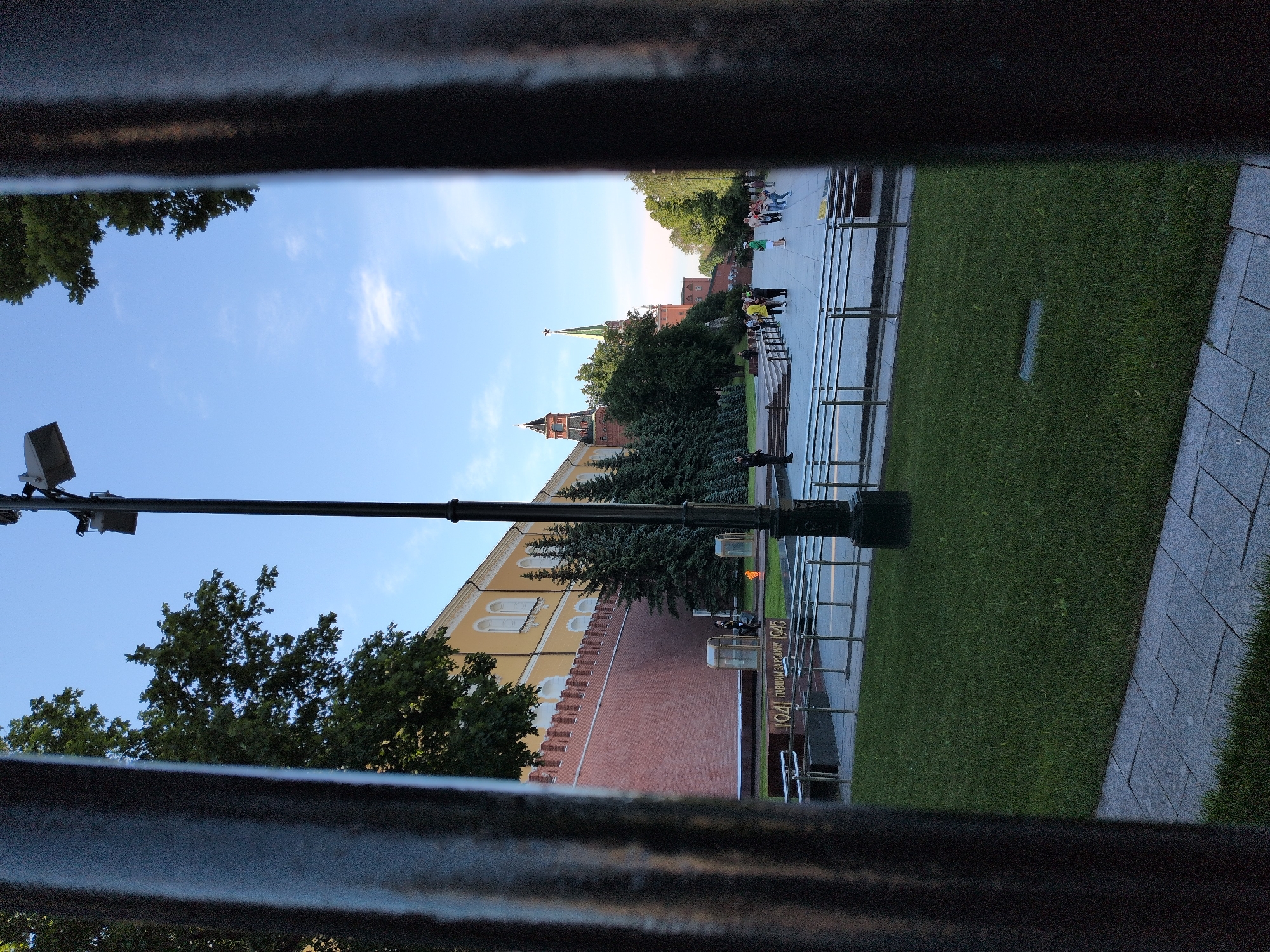 The image captures an outdoor scene, viewed horizontally through the dark, blurred bars of what appears to be a fence or railing. The bright blue sky, scattered with light white clouds, dominates the upper half of the framed view, while green grass and paved areas occupy the lower half.
In the mid-ground, a prominent red brick wall, characteristic of historical fortifications, stretches across the scene. Behind this wall, a light yellow building with arched windows and a white facade is visible. Further back and slightly to the right, a distinctive green-spired tower with a red star at its pinnacle rises above the treeline, suggesting a significant architectural landmark. Lush green trees, including both coniferous and deciduous types, dot the landscape, partially obscuring parts of the buildings.
Several people are dispersed on a wide paved pathway to the right of the wall and building. They appear to be casually walking, standing, or sitting on benches, indicating a public space or tourist attraction. A tall black lamppost with a light fixture stands vertically on the left side of the image.
On the ground near the red wall, a golden-colored monument or plaque is partially visible. The text "1941" and "ПАВШИМ ЗА РОДИНУ 1945" (To those who fell for the Homeland) is clearly discernible on this monument, implying a memorial related to World War II. A small, bright flame or light can be seen near this monument.
The scene is well-lit by daylight, indicating late morning or early afternoon with clear, sunny weather. The overall impression is one of calm, open-air sightseeing in a historically significant location.