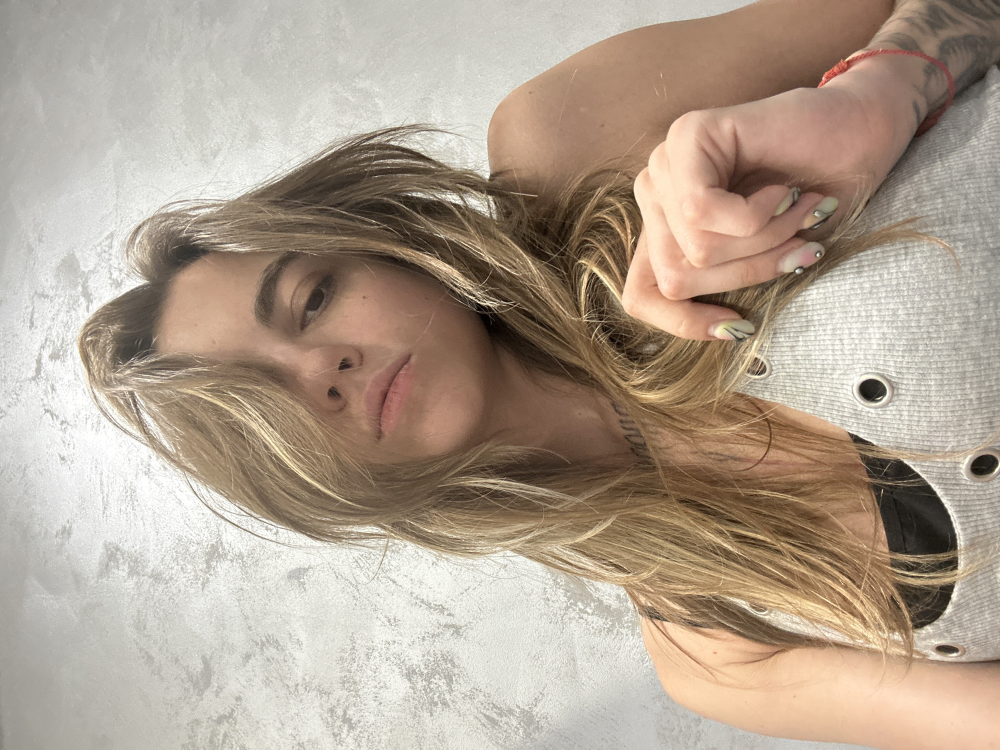 The image shows a close-up, selfie-style shot of a young woman with blonde, layered hair. She is wearing a grey tank top with a black bra underneath. The tank top has several silver grommets. Her left hand, with painted nails, is resting on her chest. A red string bracelet is visible on her wrist, and tattoos are present on her arm. The background is a textured, light-colored wall. The scene does not provide any information about the city of Moscow, Russia.