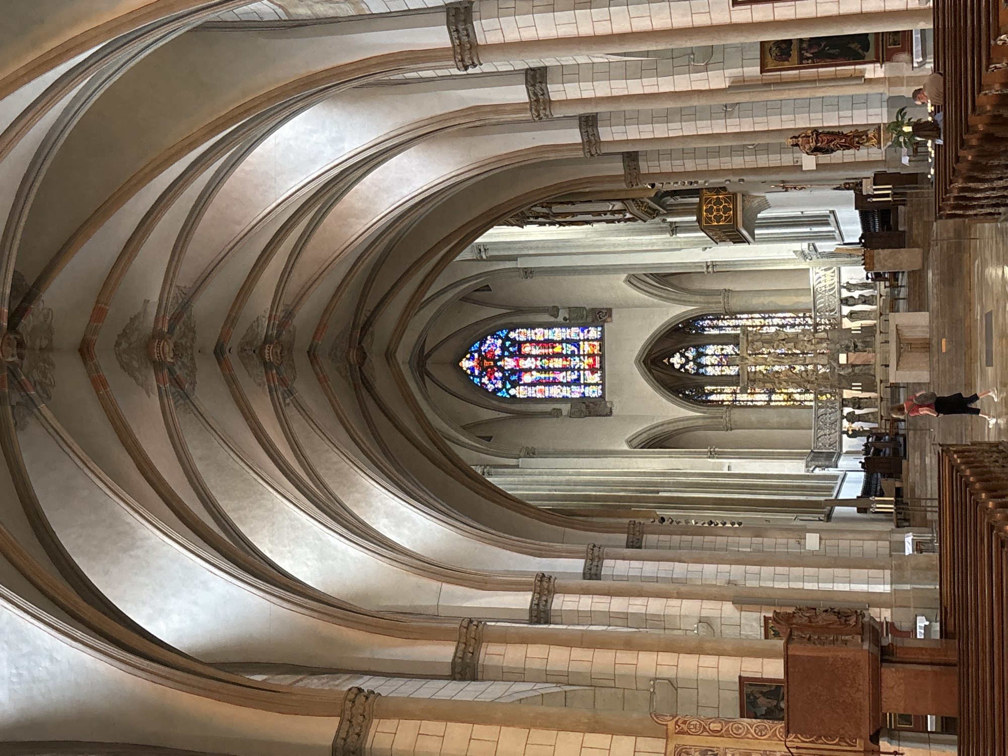 This image depicts the interior of St. Anne's Church in Augsburg, Germany. The perspective is from the nave, looking towards the altar. The architecture features soaring ribbed vaults and pointed arches, characteristic of Gothic style. Light streams through large stained-glass windows, notably a prominent rose window at the far end and lancet windows to its sides, illuminating the space with colorful hues.
Several architectural elements are visible:
- massive stone columns with decorative capitals
- wooden pews arranged in rows filling the nave
- a raised pulpit on the left side of the nave
- a choir area with a central altar and ornate screens or structures behind it
In the foreground, a person with red hair, wearing a pink shirt and black pants, is walking away from the camera towards the altar. They have a backpack. In the middle ground, to the right, two individuals are seated in the pews, facing away from the camera. To the left, a person in a red shirt and black pants is walking towards the altar.
The overall atmosphere is one of grandeur and historical significance, typical of a European cathedral.
