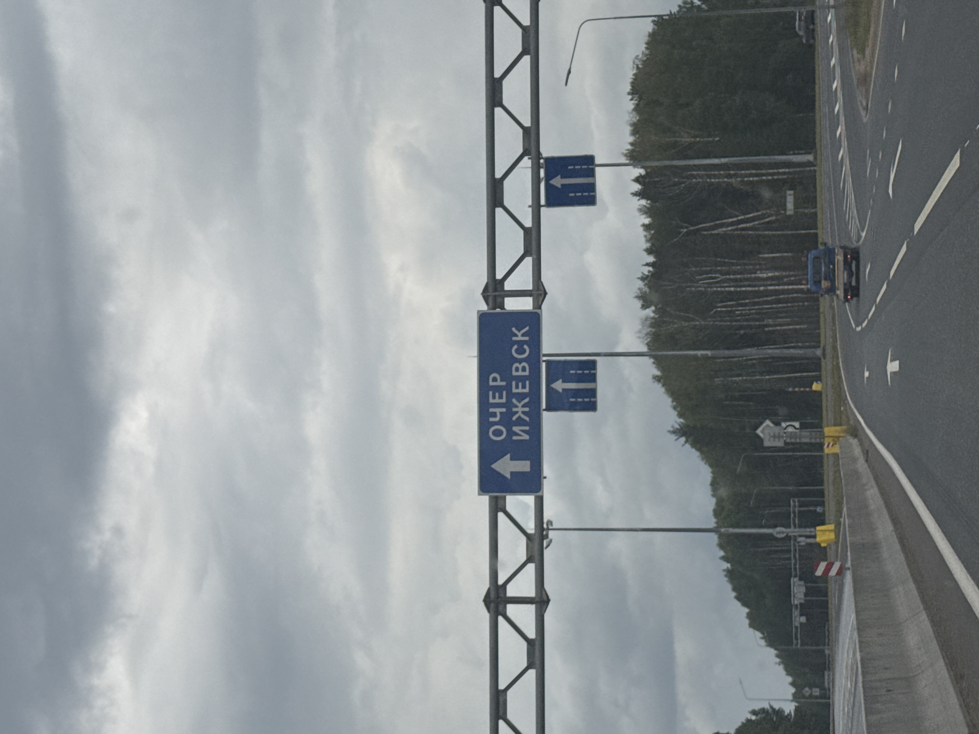 This is a photograph taken from a vehicle on a road. A blue sign overhead displays Russian text "ОЧЕР ИЖЕВ" with an upward arrow, indicating directions to Cher and Izhevsk. Below this, two blue signs with upward arrows are visible. The road curves to the right, with white lane markings. A small blue truck is driving ahead on the road. On the left side of the road, a concrete barrier is present. Beyond the road, a dense forest is visible under a cloudy sky. The image appears to be taken in or near Selishcha, Russia, based on the context provided.