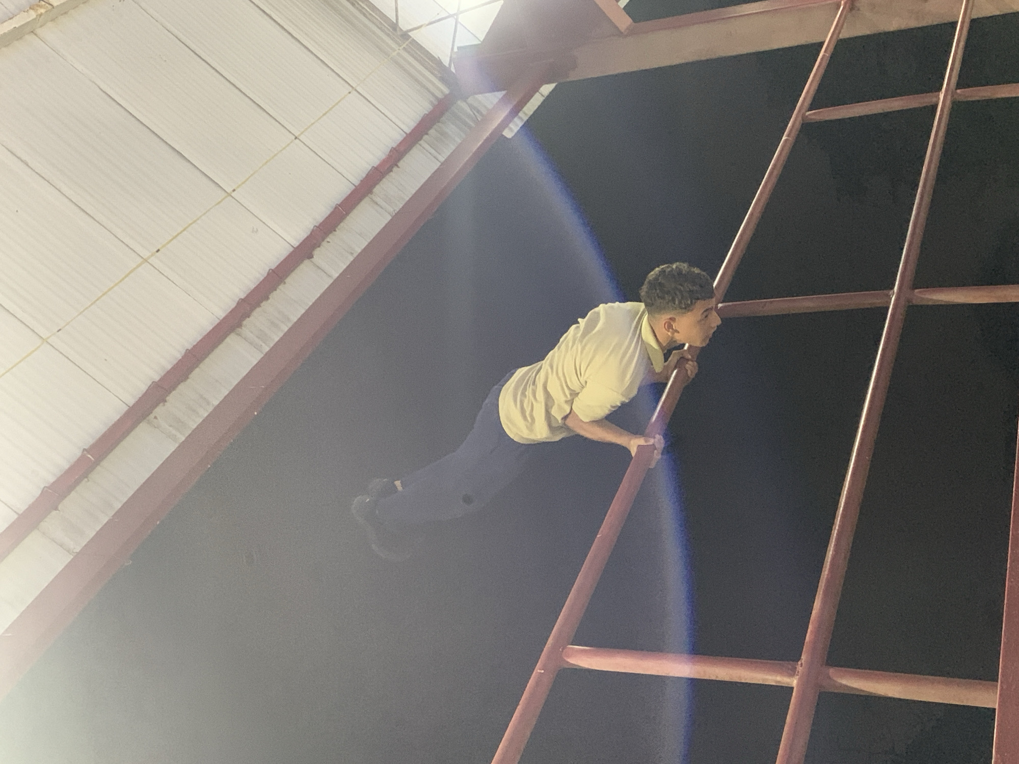 A young man is captured in a dynamic pose at night, likely indoors or in a covered outdoor area, in Santa Ana, Venezuela. The image is composed at an angle, making the man appear suspended horizontally. He is wearing a light-colored polo shirt and dark pants with sneakers, and has short, curly hair. He is holding onto a robust, red metal structure that resembles a large ladder, scaffolding, or a fixed exercise bar, with both hands. His body is held stiff and parallel to the ground or slightly angled upwards, suggesting he is performing an athletic feat or posing.
The background is predominantly dark, indicating nighttime. A bright artificial light source, possibly a spotlight or floodlight, is positioned to the upper left, casting a strong lens flare across the image, particularly evident near the man's lower body and the red structure. To the upper left, a white corrugated metal roof or ceiling structure is visible, suggesting a large building like a warehouse, gym, or covered facility. The red metal structure appears to be a permanent fixture, possibly part of the building's support or a dedicated fitness apparatus. No visible text is present in the image.