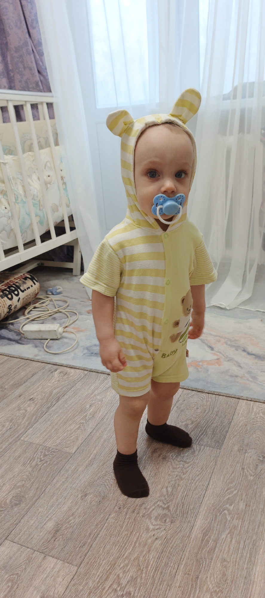 A young child stands in a room, likely a home interior, based on the presence of a crib and flooring. The child wears a yellow and white striped hooded jumpsuit with ears and brown socks. They have a blue pacifier in their mouth. The room has light-colored wooden flooring and a patterned rug. A crib with white rails and patterned bedding is visible in the background, along with white curtains and a bundle of electrical wires. The location is the city of Chaikovsky, Russia.