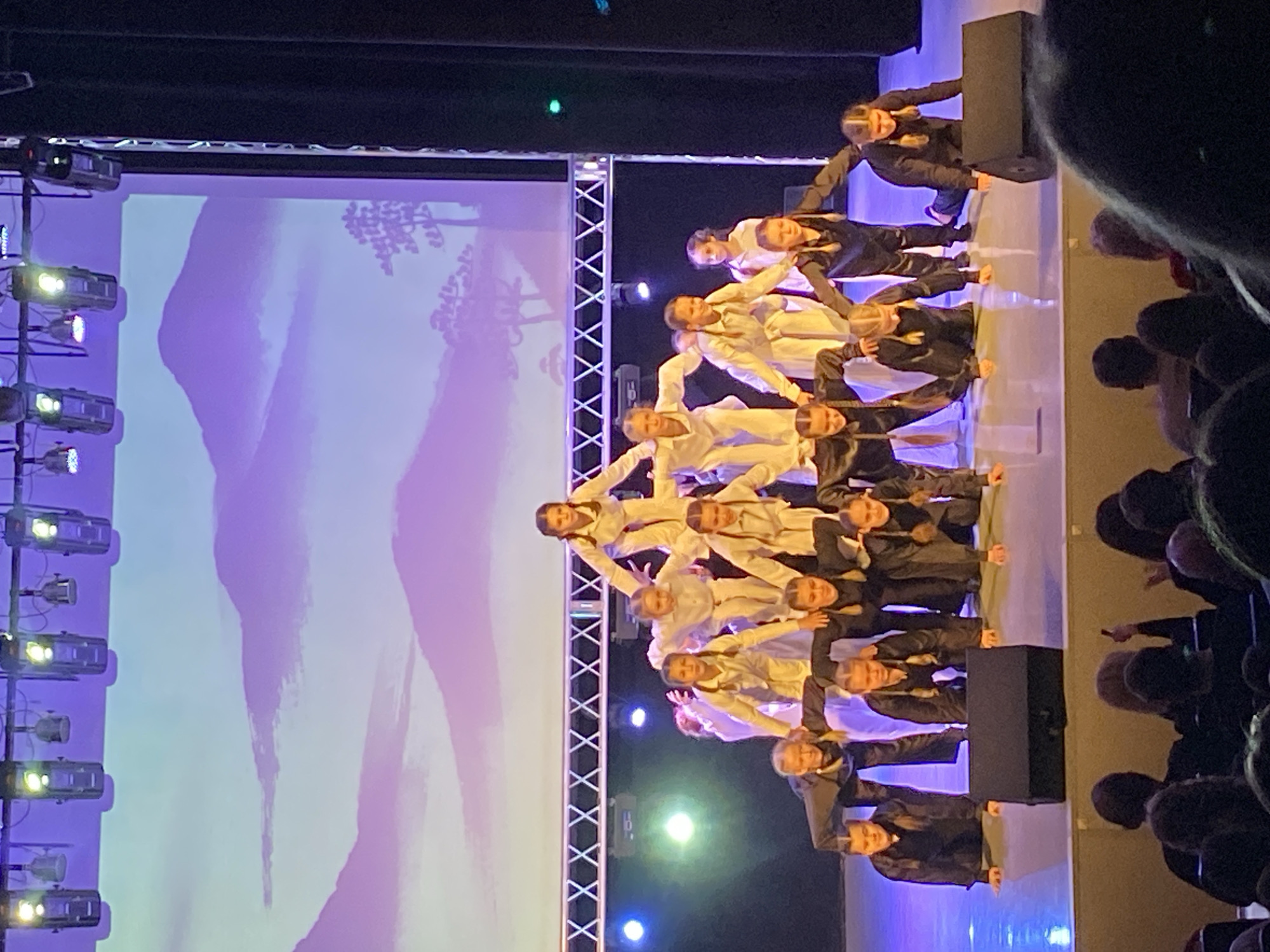 The image shows a group of children performing on a stage in what appears to be a theatrical production. The children are arranged in a pyramid formation, with some dressed in white costumes and others in black. The pyramid is illuminated by stage lighting, and a large screen in the background displays an abstract image. The photograph was taken in Chaikovsky, Russia. An audience is visible on the right. A vertical lighting rig is on the left.
