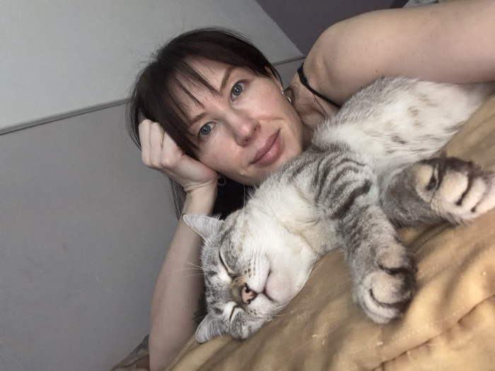 The image captures a close-up indoor scene in Perm, Russia, featuring a female individual and a domestic cat. A woman with dark brown hair, light-colored eyes, and fair skin is positioned in the upper right, looking directly at the camera with a slight smile. Her right hand is curled under her chin, supporting her head. She wears a dark-colored garment with a thin strap visible on her left shoulder. A small dark tattoo is partially visible on her left upper arm. Resting immediately adjacent to her, predominantly on the left and center, is a silver-grey tabby cat with distinct dark stripes. The cat is lying on its back, eyes closed, indicating it is asleep or resting deeply. Its front paws are curled near its chest, and its rear paws are extended. Both the woman and the cat are on a golden-brown, textured fabric surface, likely bedding. The background consists of a plain light grey wall with a darker horizontal molding or trim running across it.