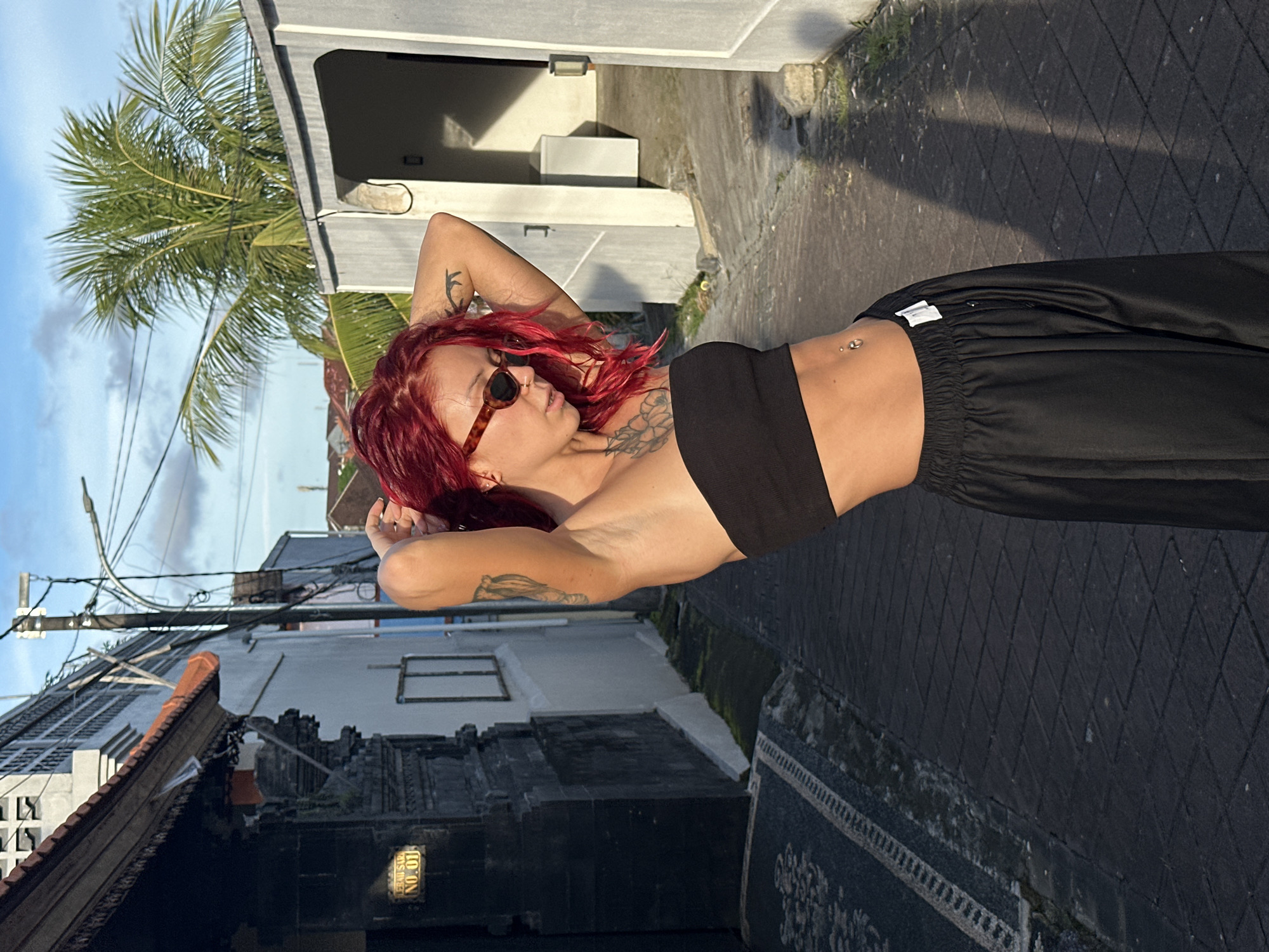 A young woman with vibrant red hair is featured prominently, posing outdoors in what appears to be a street or alleyway in a tropical location, consistent with North Kuta, Indonesia. She is wearing dark, tortoiseshell-patterned sunglasses, a black bandeau top, and loose-fitting black pants, revealing a navel piercing. Her arms are raised with hands behind her head, showcasing tattoos on both arms and her left shoulder. The woman projects a confident and relaxed demeanor.
The scene is illuminated by the warm, directional light of late afternoon or early evening, creating long, distinct shadows on the paved ground. Behind her, buildings line the street. On the left, a bright green palm tree extends towards a clear blue sky with some wispy clouds and overhead utility wires. The architecture includes a light-colored, modern-looking building and a darker, intricately carved structure, possibly a traditional Balinese building or temple, near the lower left. On this darker building, a small sign is visible with the text "NO. 01", with other partially obscured text beneath it. The paved ground shows a diamond pattern, and a decorative pattern resembling a rug or intricate paving is also present on the ground to her right. The overall atmosphere is warm and serene.