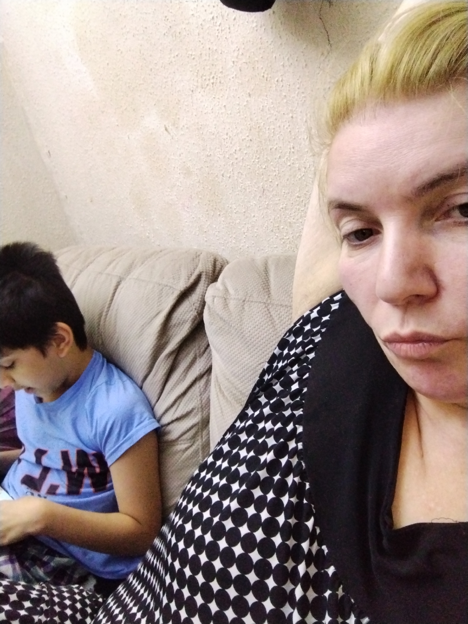 A woman with blonde hair sits on a beige couch, looking toward the camera with a neutral expression. To her left, a young boy with dark hair is seated, looking down at something in his hands. The woman wears a black top with a white polka dot pattern on the front. The boy wears a blue shirt with black lettering. The background is a light beige wall. The location is unknown and cannot be determined from the image.