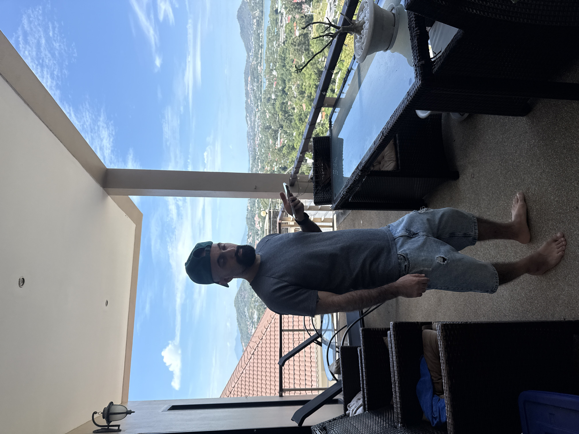 A man with a beard and a green baseball cap is standing on a balcony in Ko Samui District, Thailand. He is wearing a dark gray t-shirt and ripped denim shorts. He is holding a mobile phone in his right hand and looking down at it. In the background, there is a view of lush green hills, buildings, and a body of water. The balcony is furnished with wicker chairs and a glass-topped table. There are also lounge chairs and a potted plant on the balcony. The sky is blue with some white clouds.