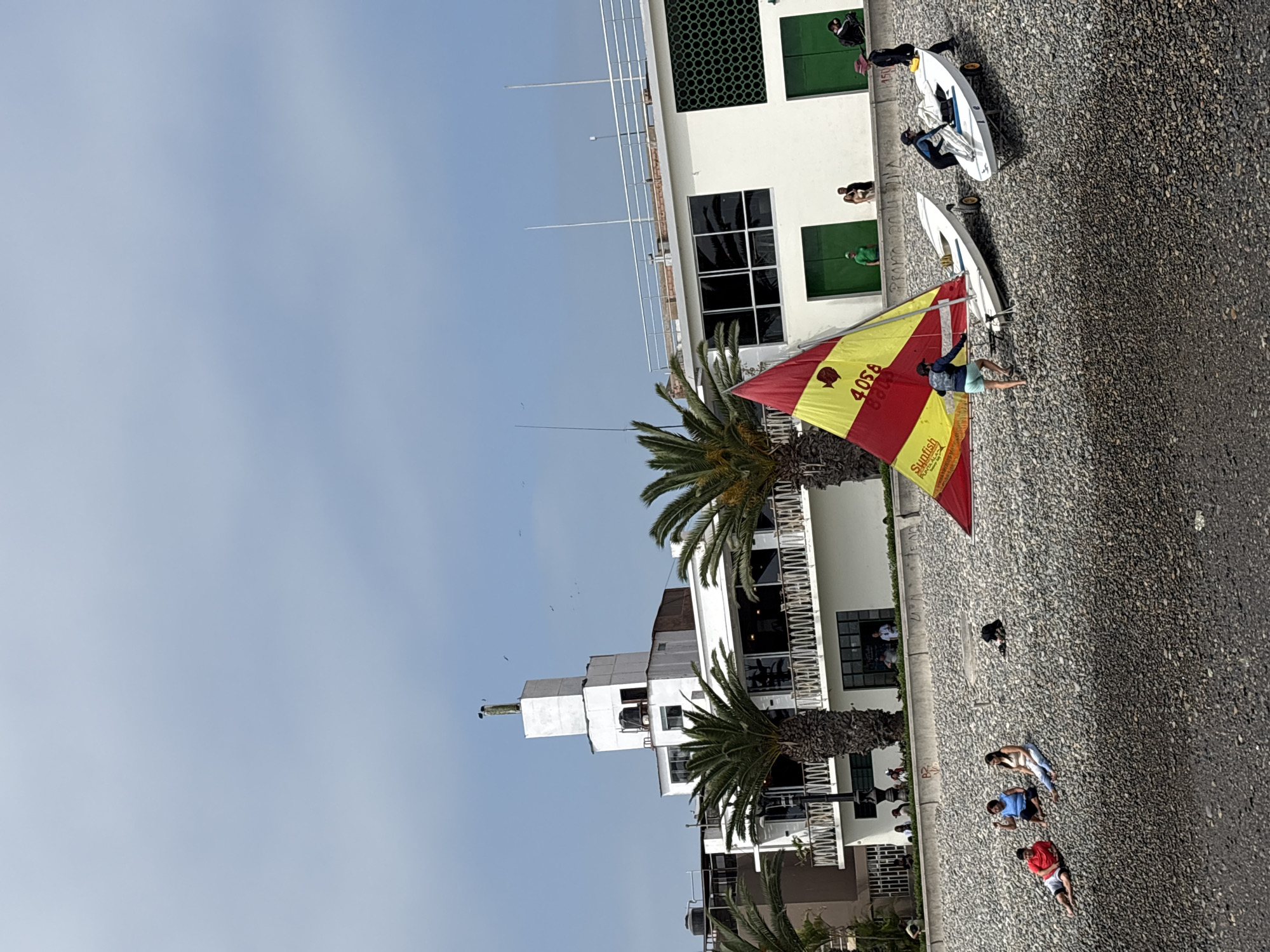 The image depicts an outdoor scene in La Punta, Peru. A Sunfish sailboat with a red and yellow sail is being prepared for sailing by a person wearing shorts and a jacket. There are two additional sailboats nearby, lying on what appears to be a paved or pebbled ground next to a white building. A group of people are lying on the ground further away. Palm trees are present. The sky is light blue with few clouds.