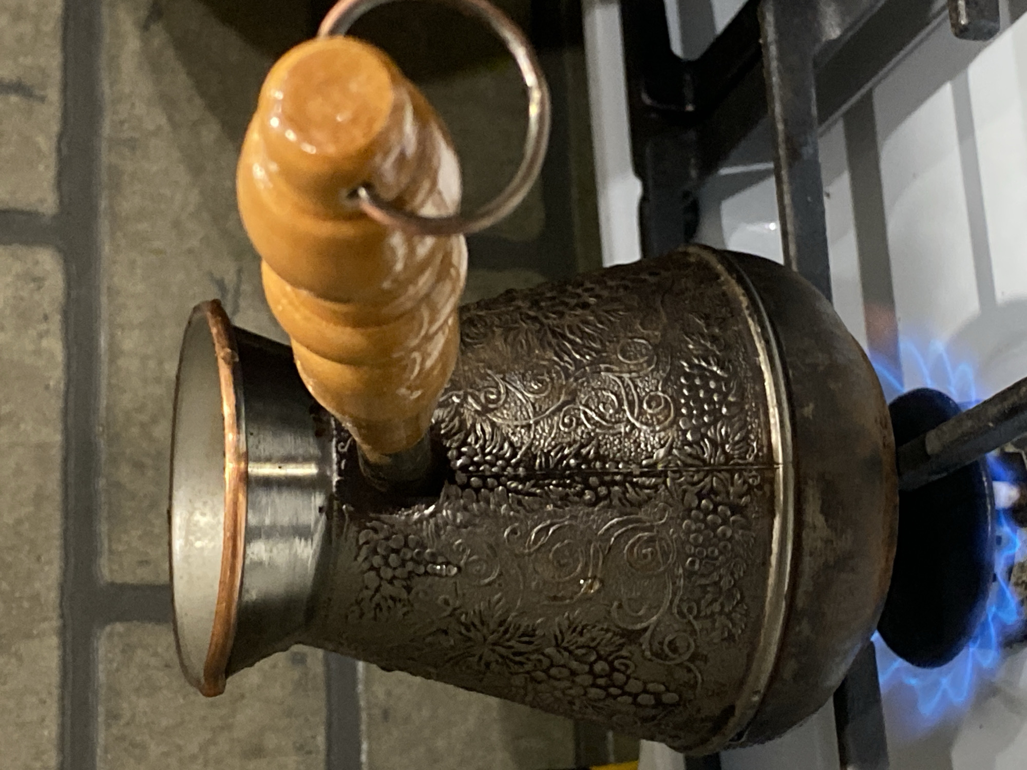A metallic coffee pot with a wooden handle sits on a lit gas burner. The pot is decorated with embossed designs. The background includes a stove and tiled surfaces. There is no evidence in the image to suggest its location is Sochi, Russia.