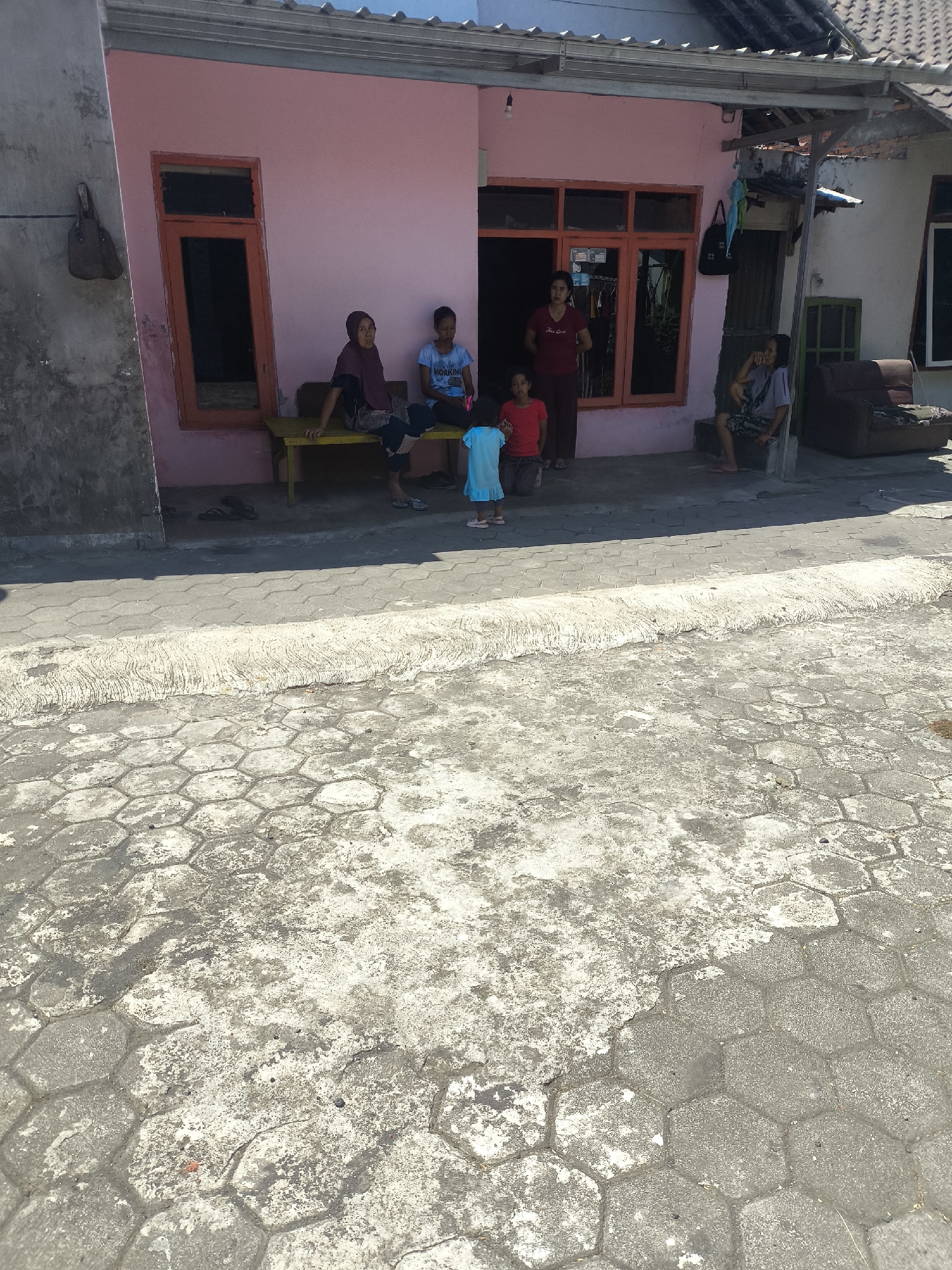 In front of a pink residential building in Kebonsari, Indonesia, a group of people are gathered on the sidewalk and porch. A young child in a blue dress stands on the patterned concrete sidewalk, facing a young girl in a red shirt. To their right, on a yellow bench, sit two women, one wearing a dark headscarf and the other a light blue t-shirt. Further to the right, a woman in a red t-shirt stands by the doorway of the building, which has orange-framed windows. Next to her, on a low step, a person wearing a patterned t-shirt and dark pants is seated, with a dark sofa visible behind them. The building's exterior is a vibrant pink, and the surrounding area is paved with hexagonal concrete blocks.