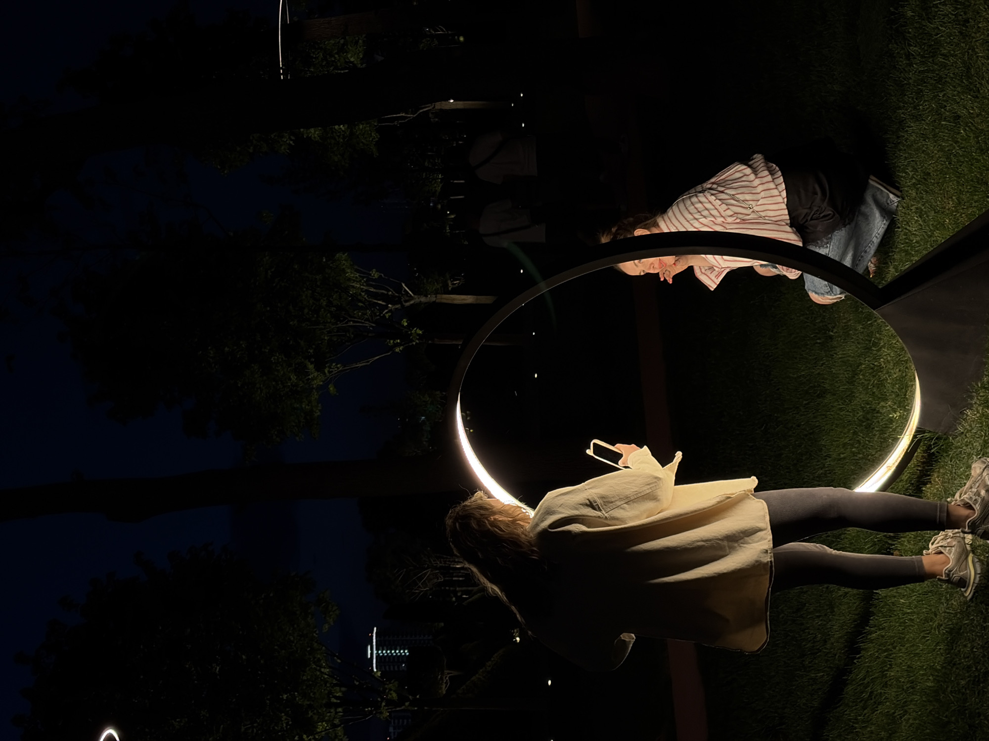 At night, two women are standing and looking at each other through a circular mirror frame in a park in Krasnodar, Russia. The woman facing the camera has blonde hair and is wearing a beige coat, leggings, and athletic shoes, and holding a cellphone in her hand. She is standing on green grass. The other woman is wearing a red and white striped shirt and jeans. The park also has trees and tall, thin lights. In the background, there is a building with windows illuminated.