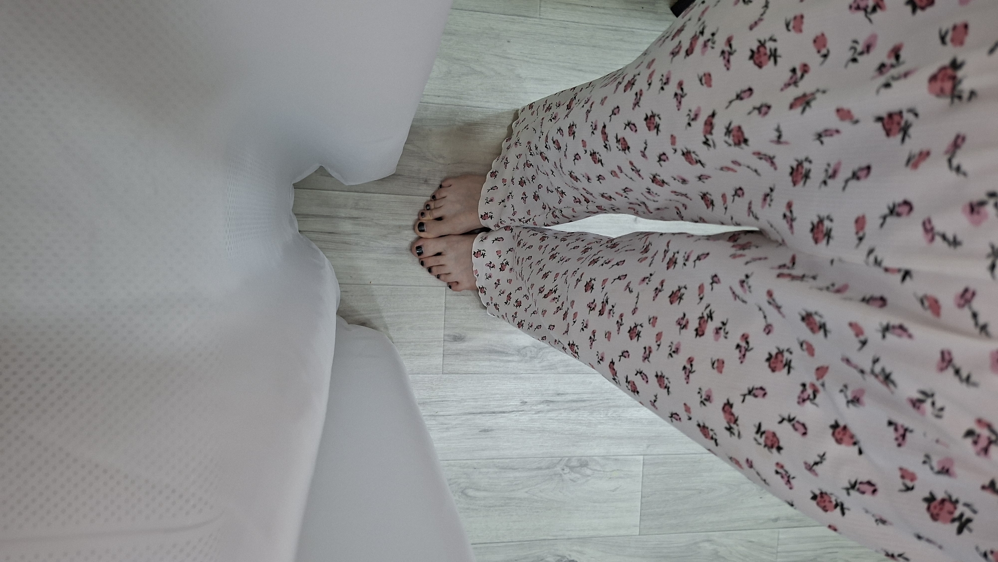 The image shows a close-up, overhead view of a person's legs and feet. The person is wearing light-colored pajama pants with a floral print of small pink and red flowers. Their feet are bare, with dark nail polish on their toenails. The feet are positioned on a light-colored wooden floor. To the left of the person's legs and feet is a large, soft, white object, possibly a sofa or a bed, with a textured surface. The scene appears to be indoors. The lighting suggests it could be daytime, given the natural light implied by the scene. There is no visible text in the image. The overall impression is a casual and relaxed domestic setting.