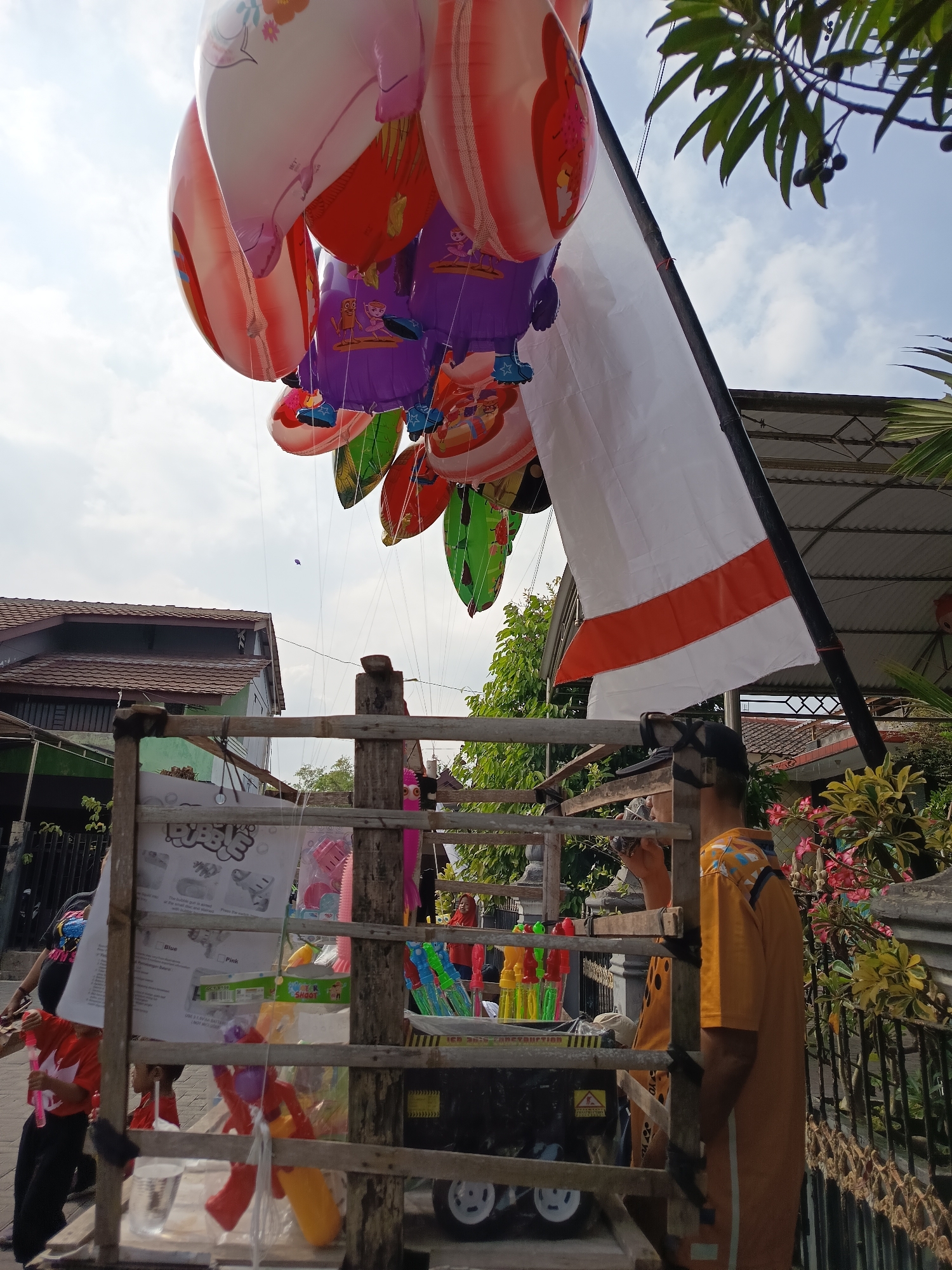 In Ngaglik, Indonesia, on a bright, cloudy day, a street vendor is selling balloons and bubble toys from a wooden cart. The cart displays various colorful toys, including bubble guns and wands, alongside a sign for "Bubble Toy." A cluster of helium balloons, predominantly red, white, and purple, floats above the cart, some depicting cartoon characters. A white flag with an orange stripe is also attached to a pole on the cart. Two young boys in red shirts are visible, one holding a pink bubble wand, near the cart. In the background, residential buildings with tiled roofs and lush green foliage suggest a suburban or rural setting. A man wearing an orange shirt and a cap stands near the cart, appearing to be the vendor. The scene is captured from a low-angle perspective, looking upwards towards the balloons and the sky.