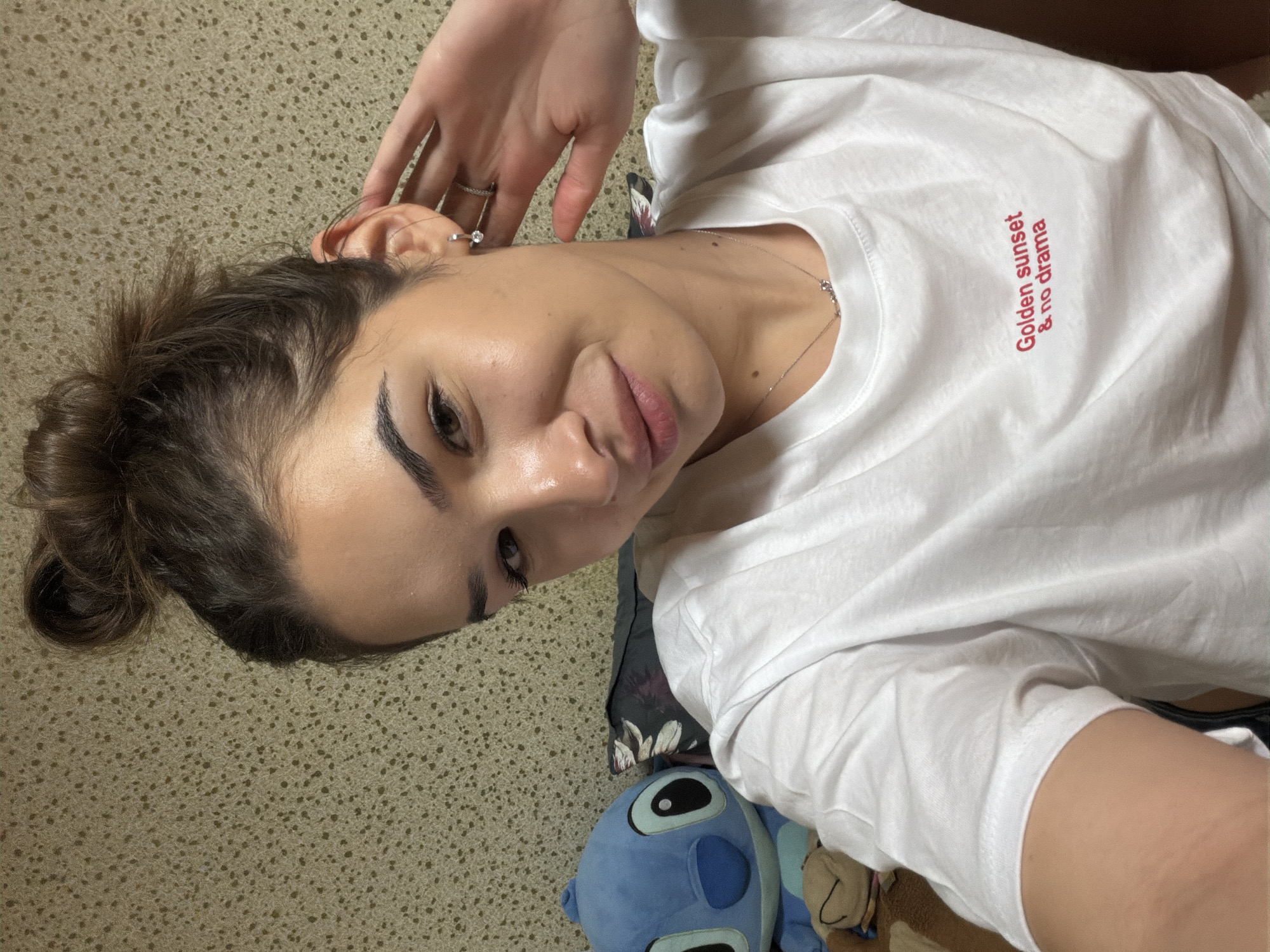 A woman with brown hair styled in a bun is lying down on a surface with beige and brown speckled carpet. She is wearing a white t-shirt with the phrase "Golden sunset & no drama" printed in red text on the upper right side. She has on earrings and a delicate necklace. Her right hand is raised near her ear. Next to her is a blue plush toy that resembles the character Stitch from the Disney movie "Lilo & Stitch." The location is not evident from the image.