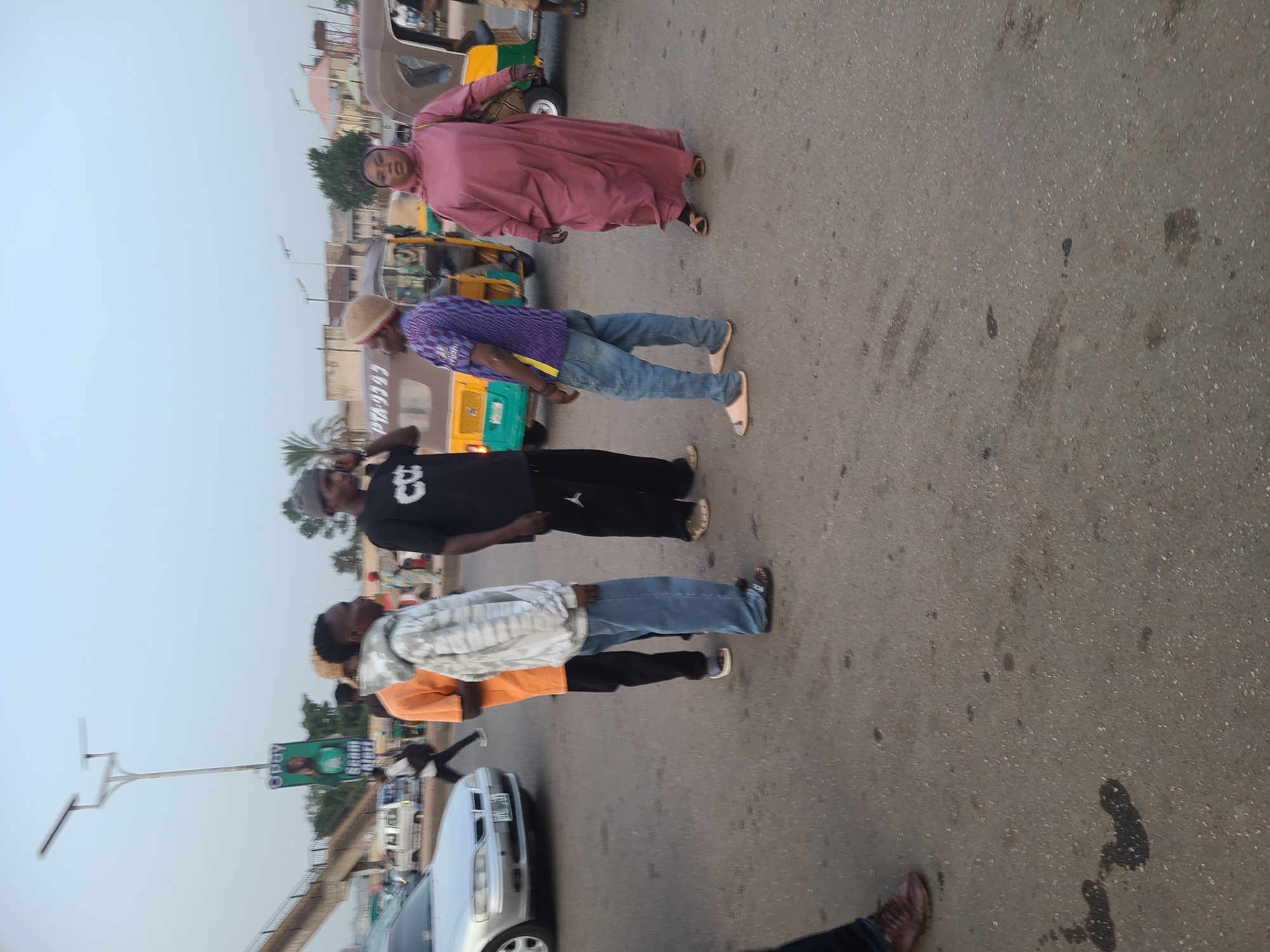 The image captures an outdoor scene in Jos, Nigeria. The location appears to be a busy street with pedestrians and vehicular traffic.
Four people are walking along the road. An elderly woman is dressed in a pink robe. A man behind her is wearing a purple shirt, jeans, and a hat. Another man is wearing a black T-shirt with a white number "8" on the back and black pants. The man in front of him is wearing a white patterned shirt, an orange shirt, and blue jeans.
A silver car is parked on the side of the road. Yellow tricycle taxis are in the background.
A light pole and overhead bridge are also visible in the image. The sky is overcast, and the overall lighting suggests daytime.