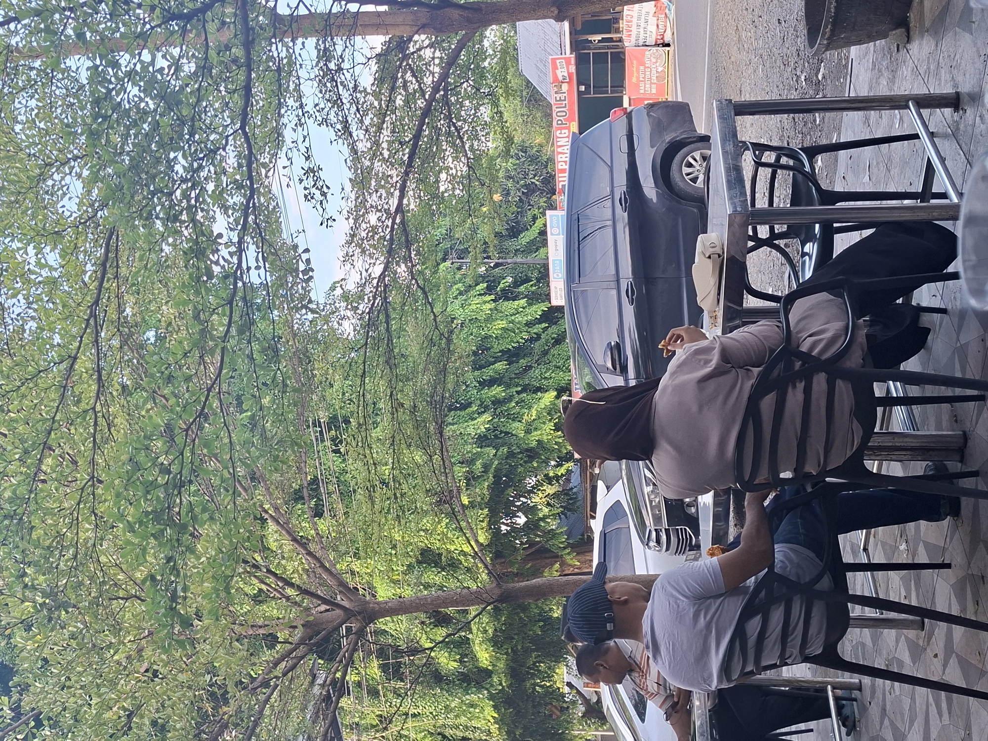 In Keuniree, Indonesia, a scene unfolds at an outdoor dining area adjacent to a road. Three individuals are visible, two seated at a metal table and one standing to the left. A man wearing a gray t-shirt and a blue and white striped cap is seated, facing away from the camera. He is wearing blue jeans. To his right, a person in a beige hijab and a light brown cardigan is also seated, facing away, with a small white purse on the table. A third individual, partially visible on the left, wears a striped shirt and is looking at a phone. On the table, there are some food items and a small white purse. In the background, a black SUV is parked on the road, along with several other vehicles. Behind the vehicles, a building with signage is visible, including advertisements for services like "DANA" and "GOPAY," and a sign that reads "IL PRANG POLEN." Trees with green foliage overhang the dining area, and a power line runs across the scene. The ground is paved with geometric tiles.