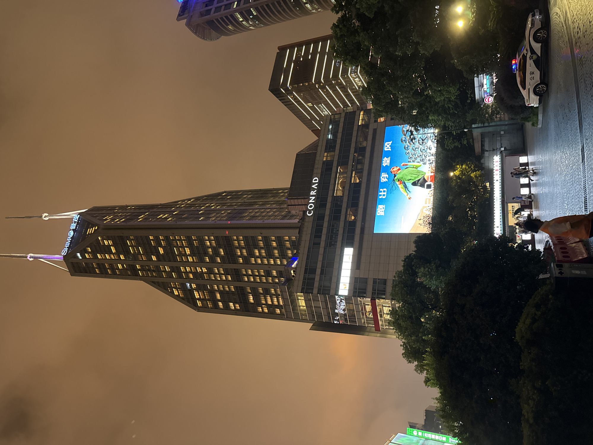 This is a night photograph taken in Shanghai, China, likely in the Lujiazui financial district. The primary subject is a modern skyscraper with a distinctive angular design, illuminated by numerous windows. The building's upper section features a neon sign that reads "SHIMAO" in English, with Chinese characters next to it. Below this, the building's facade displays the "CONRAD" hotel logo.
A large digital billboard is mounted on the lower section of the skyscraper, displaying a vibrant advertisement featuring a person in athletic wear running against a backdrop of circular motifs. The billboard is brightly lit, contrasting with the dimmer surroundings.
In the foreground, several trees with lush foliage obscure part of the scene. On the right side of the image, a white police car with flashing red and blue lights is parked. The police car has "公安" (Gōng'ān), meaning public security, written on its side. There are a few individuals on a paved walkway in front of a building structure with multiple entrances. The overall atmosphere is urban and dynamic, typical of a bustling city at night. The sky is overcast and tinged with a warm orange glow, likely from city lights and atmospheric conditions.