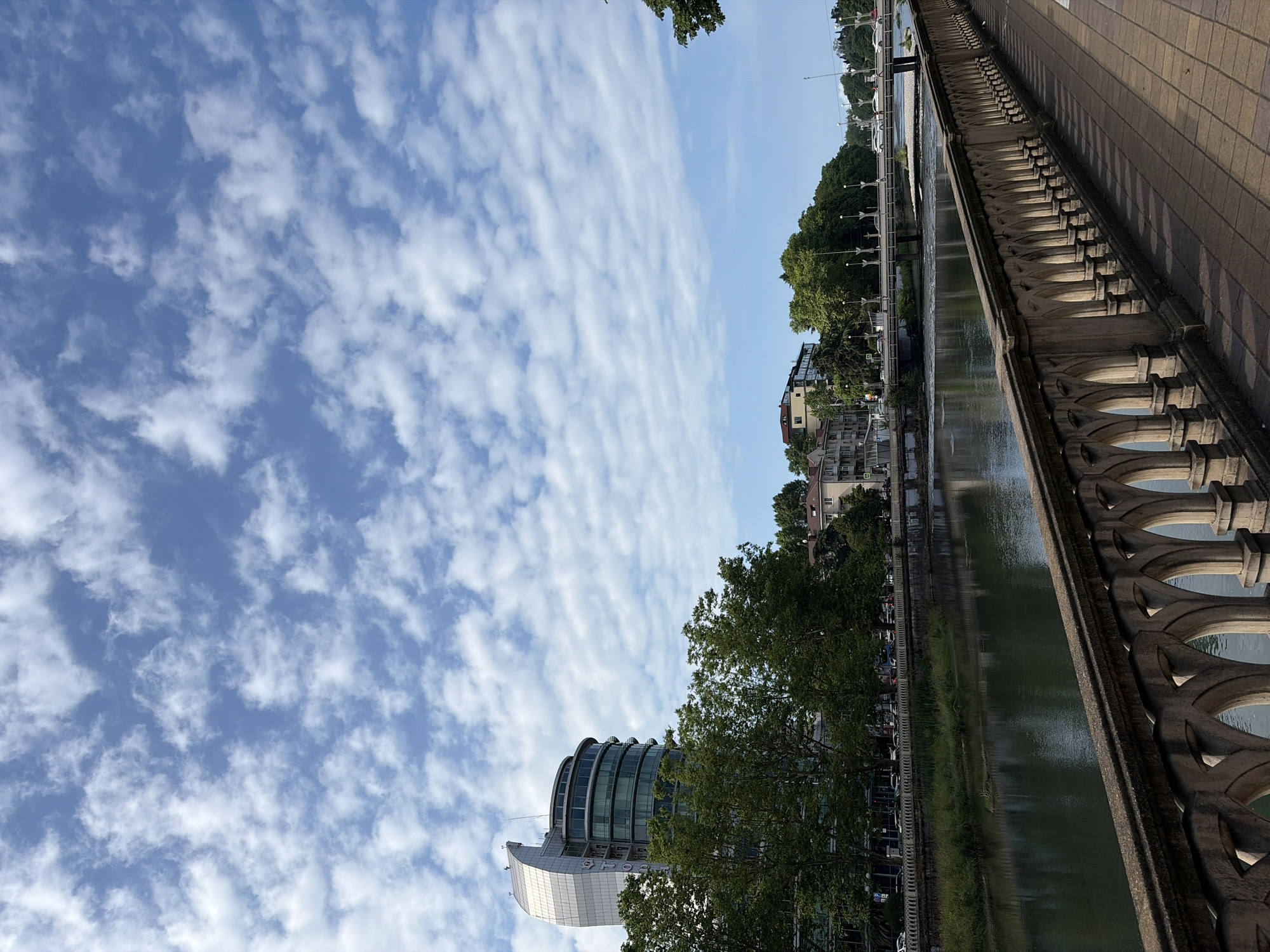 The image shows a scenic view of the city of Sochi, Russia, likely along the Matsesta River. In the foreground, a decorative stone balustrade with arched openings runs along a paved walkway. The walkway is made of brick pavers, with shadows cast by the balustrade. To the left of the walkway and balustrade, the Matsesta River flows gently, with its surface reflecting the sky and surrounding environment. On the opposite bank of the river, a row of trees with lush green foliage lines the embankment. Behind the trees, several buildings are visible, including a modern, curved glass-fronted building on the left and other multi-story buildings further down the river. A bridge with traffic, including cars, spans the river in the middle ground. The sky above is a bright blue, filled with scattered white cumulus clouds. The overall scene depicts a tranquil urban riverside setting. There are no visible people in the image.