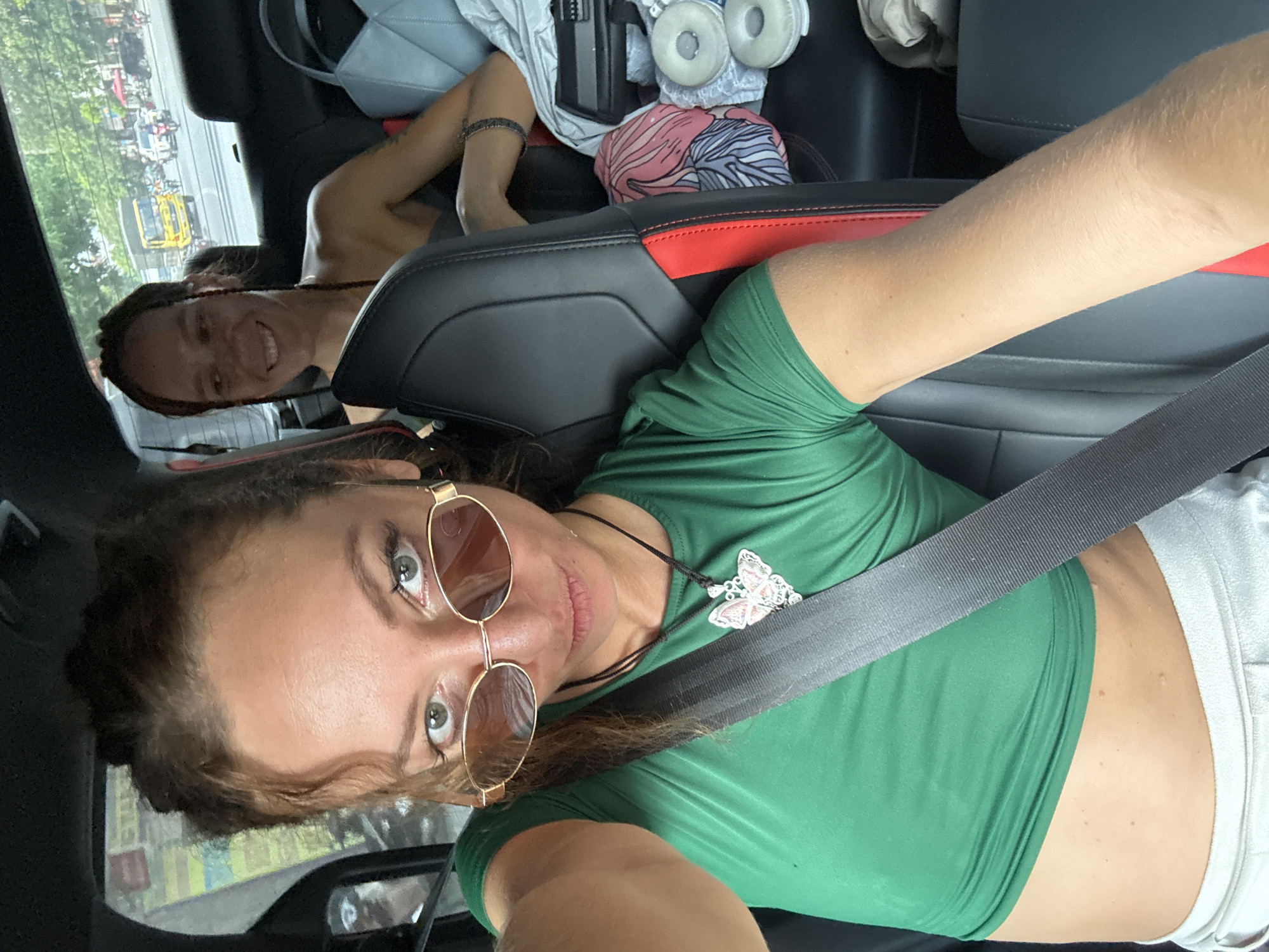A selfie taken inside a car captures two women. In the foreground, a woman with light brown, curly hair, wearing amber-tinted sunglasses, looks directly at the camera with a calm expression. She wears a green crop top, exposing her midriff, and light-colored bottoms. A black seatbelt crosses her chest diagonally, and she has a butterfly pendant necklace. Her left arm is extended to take the photo, with her elbow resting on the car's black and red accented seat.
In the background, in the back seat, another woman with dark hair, possibly in dreadlocks or braids, smiles broadly at the camera. The car's interior features black leather seats with red detailing. Various items are visible on the back seat, including white headphones, a light grey bag, and a patterned fabric item.
Through the car window, a bright daytime street scene unfolds. Green trees line the road, and multiple vehicles, including motorcycles and a distinct yellow truck or bus, are visible, suggesting a busy thoroughfare. The setting is inside a moving or stopped vehicle, likely in Hà Giang 1 Ward, Vietnam. The time of day is clearly daytime, indicated by the bright outdoor lighting. No discernible text is visible.