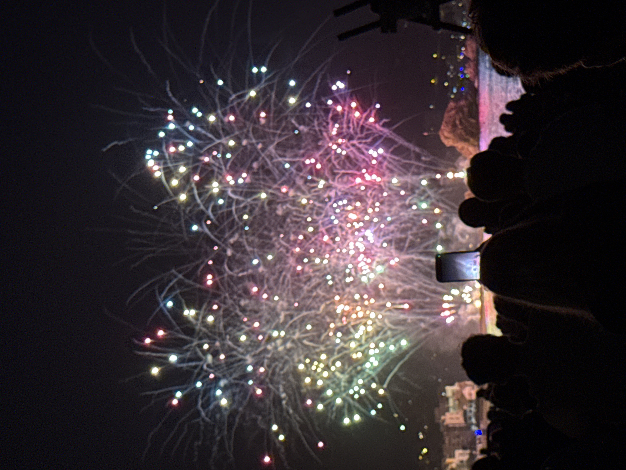 The image captures a nighttime fireworks display over the sea in Blanes, Spain, likely during the Sant Joan festival. A crowd of silhouetted people watches the colorful explosions in the sky. In the foreground, a person in the crowd holds up a smartphone, presumably to record the event. Buildings are visible in the background along the coastline. The scene is illuminated by the bright bursts of fireworks, which cast a vibrant glow on the water.