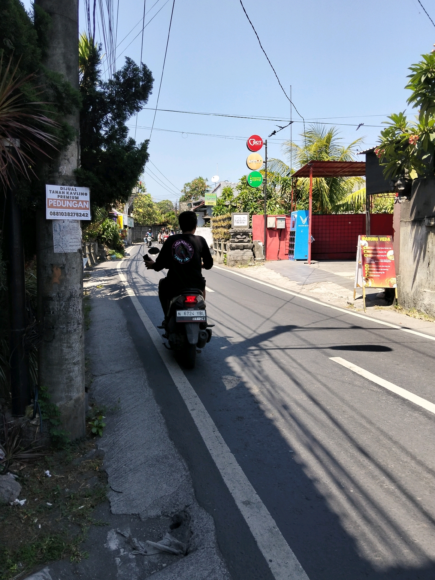 A man wearing a black t-shirt and black pants rides a black motorcycle down a paved road in Denpasar, Indonesia. The man is facing away from the camera, and the motorcycle is slightly angled to the left. The road is lined with trees and buildings on both sides, and there are power lines overhead. The sky is clear and blue. A sign on a pole to the left of the road reads "DIJUAL TANAH KAVLING PREMIUM PEDUNGAN 0881038275242". A menu sign to the right of the road reads "WARUNG VEDA". There are shadows of power lines cast on the road, indicating a sunny day.