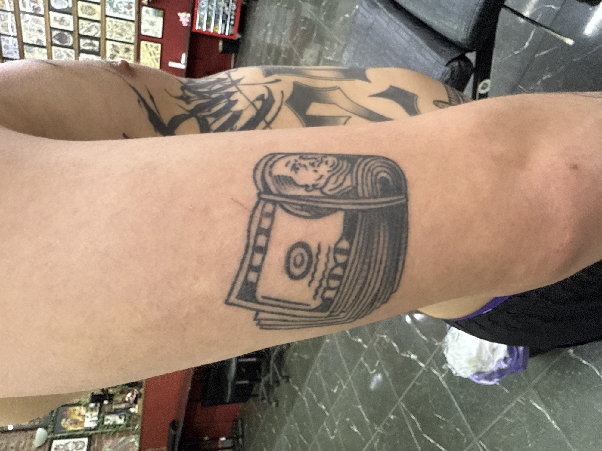 The image shows a person's arm with two tattoos. One tattoo is a stack of US dollar bills, and the other is a larger tattoo with abstract patterns. The person is seated, likely in a tattoo parlor, given the presence of tattoo flash sheets on the wall. The floor has a tile pattern, and a tattoo chair is visible in the background. The location is not specified and Moscow, Russia cannot be confirmed from the image.