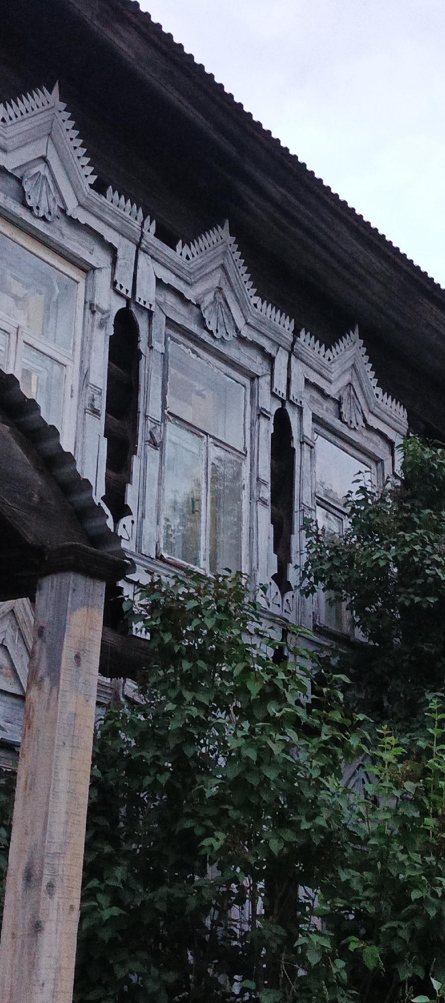 The image shows a close-up view of the upper section of a weathered wooden building with ornate decorative elements above a row of windows. The architecture suggests a traditional Russian style, possibly from Votkinsk, Russia, a city known for its historical wooden architecture. The building's facade features intricately carved wooden trim, painted a faded greyish-blue, which is peeling in places, indicating age and exposure to the elements. The roofline is visible at the top, with dark wooden shingles. The windows are multi-paned and appear to be old. Lush green foliage, likely trees or large bushes, partially obscures the lower part of the building and the windows, suggesting an overgrown or natural setting. The lighting is subdued, indicating an overcast day or evening. There are no people or animals visible in the image. The overall impression is one of rustic charm and historical character, with a sense of decay and natural reclamation.