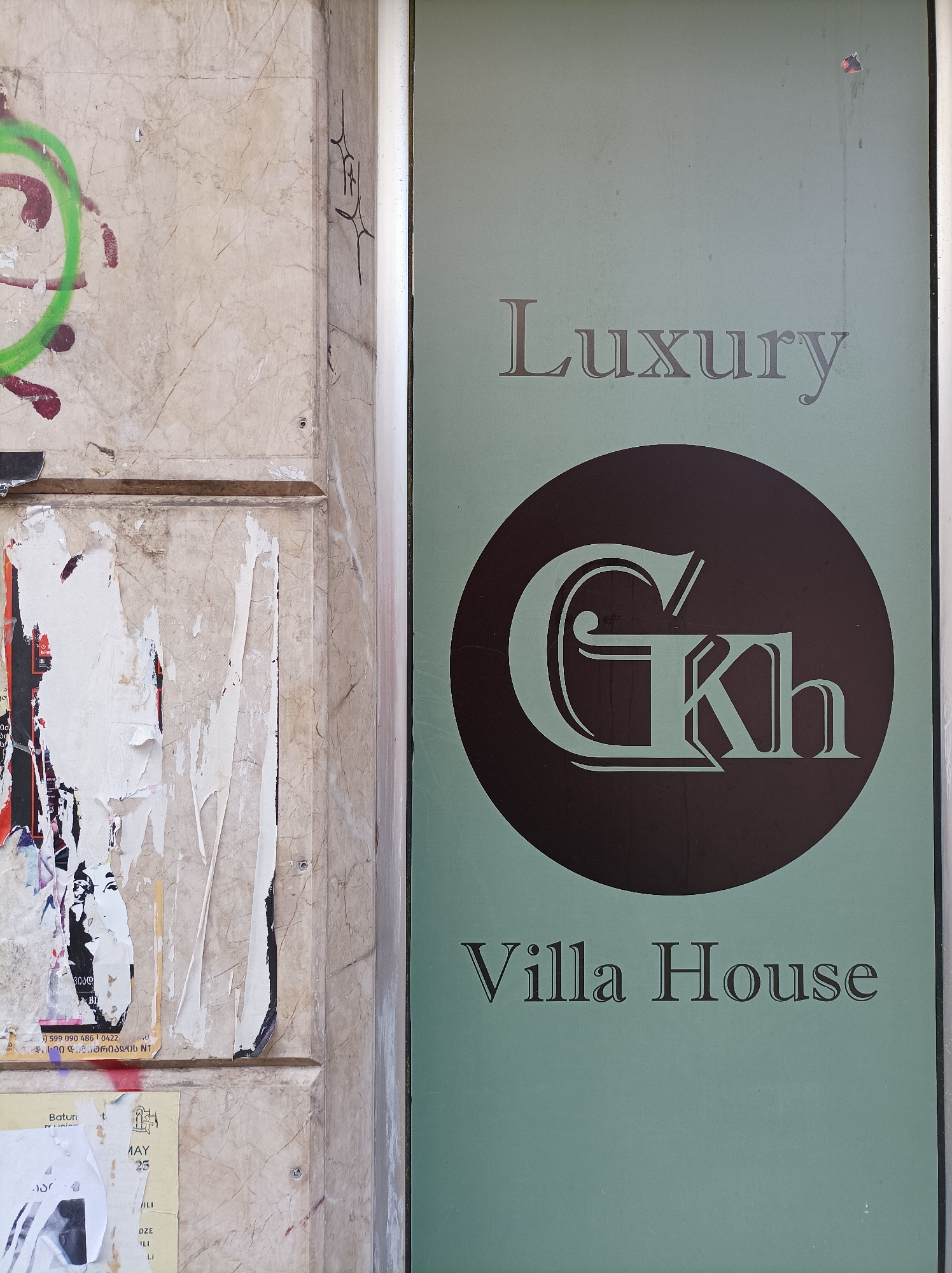 The image captures a section of a building in Batumi, Georgia, featuring two distinct areas: one is a distressed exterior wall covered with graffiti and layers of torn posters, the other is a sign for "Luxury Gkh Villa House." The sign is set against a light-green background with the words "Luxury" and "Villa House" in a dark-gray font. A stylized "Gkh" logo is prominently displayed within a black circle. The adjacent exterior wall is marred by green and red graffiti, along with remnants of posters that appear to have been repeatedly torn, revealing snippets of text in Georgian.