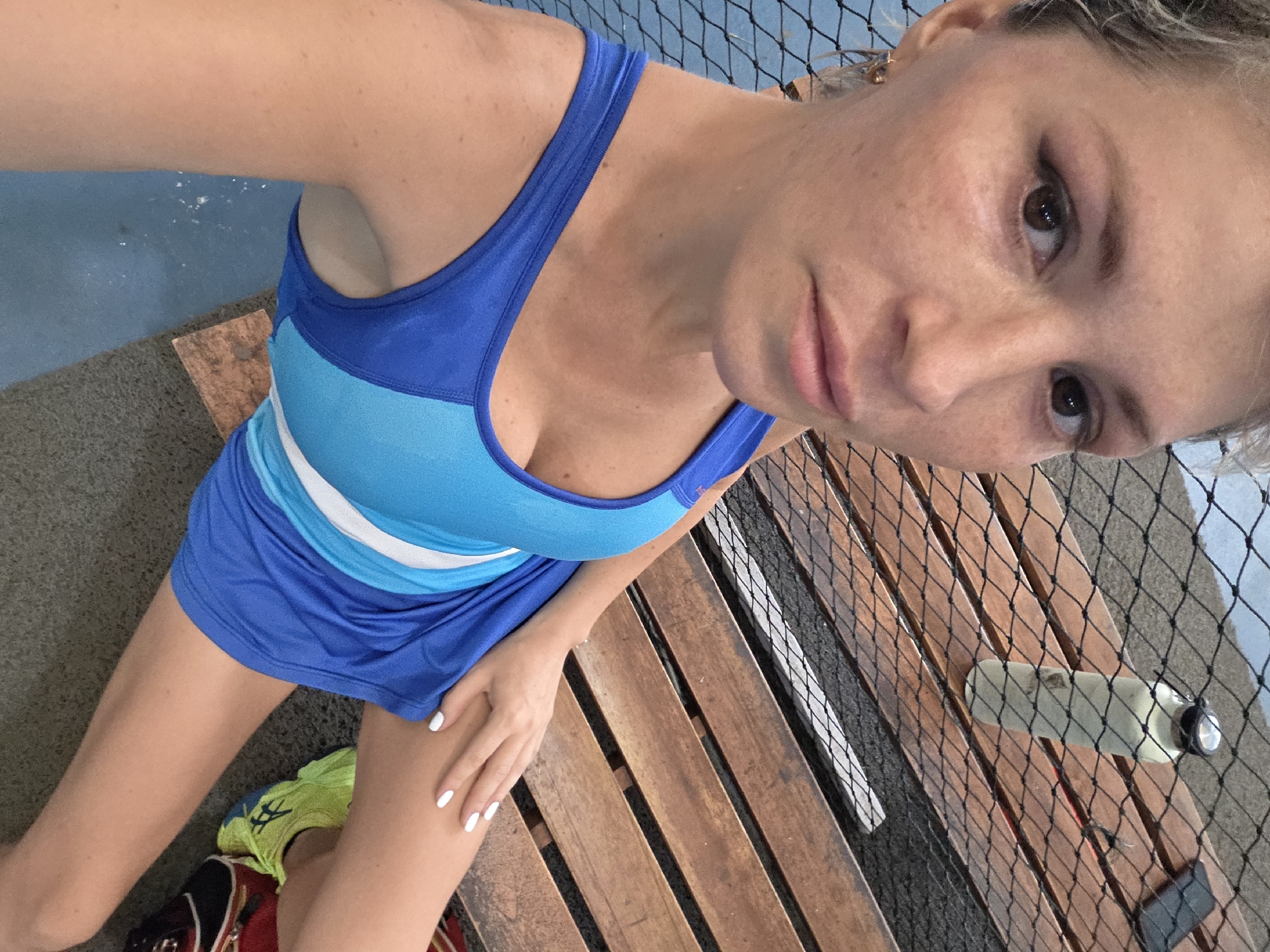 The media file shows a woman in a blue and white athletic dress, likely a tennis outfit, posing for a selfie. She is sitting on a wooden bench. Her hair is blonde, and she is wearing makeup. Her nails are painted white. To her right, a water bottle is visible on the bench, and behind her is a black net, suggesting she may be on a tennis court or a similar sports facility. The lighting indicates it is daytime. The ground appears to be concrete. A small backpack with some clothing spilling out is visible near her feet. The overall impression is that of a post-activity or pre-activity snapshot in a sports setting.