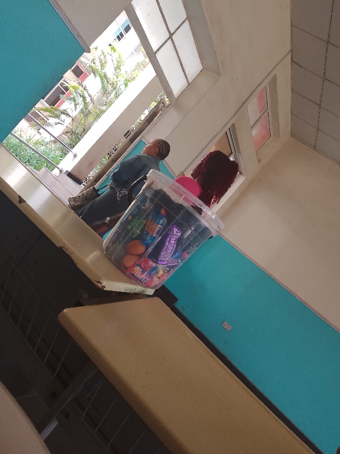A medium-shot, high-angle photograph depicts an indoor scene. Two individuals, one with reddish-brown hair, are visible in the background by a window. One person has short hair styled in braids. In the foreground, a clear plastic container filled with various snack items, including eggs, candy, and chocolate bars, rests on a surface. The context of a city is inferred by the prompt and implied through the appearance of an urban setting outside the window. The location is Umueshi, Nigeria.