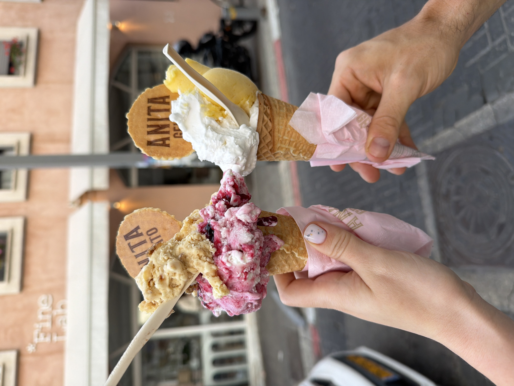 Two people are holding ice cream cones in front of the Anita Gelato shop in Tel-Aviv, Israel. The person on the right has fair skin and is holding an ice cream cone with yellow ice cream, whipped cream, a wooden spoon, and an "Anita" cookie. The cone is wrapped in a pink napkin. The person on the left has fair skin with a polka-dotted manicure, and is holding an ice cream cone with tan and pink ice cream, a wooden spoon, and an "Anita" cookie. The cone is wrapped in a pink napkin. The shop behind them is peach-colored with white trim and windows. A motorcycle is parked in front of the shop, and part of a white car is visible at the bottom of the image. The street is gray and bricked.