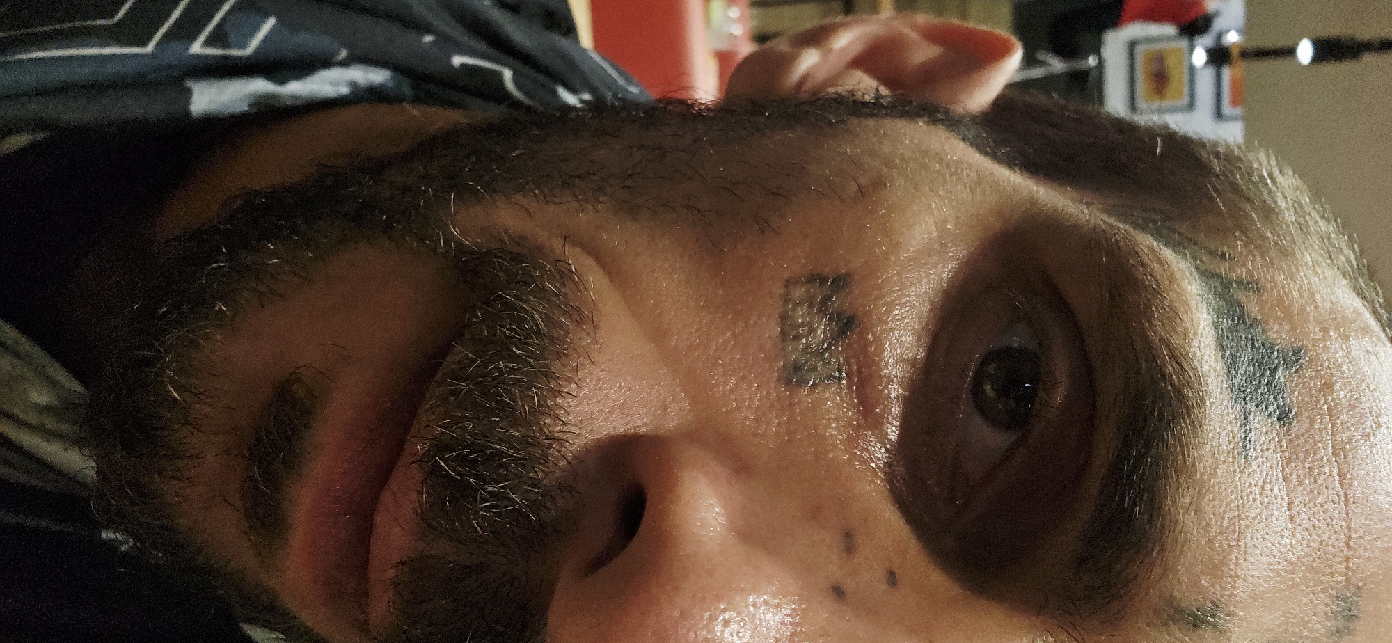 A close-up image features a man's face, tilted slightly to the left. His complexion appears light, possibly due to lighting, and his skin looks somewhat shiny. The man has dark, short beard, mustache. Distinct tattoos are visible on his forehead: one between his eyebrows that look like the letter "E" and another extending from just above his left eyebrow towards his temple. There are also what appear to be a grouping of small dark moles or blemishes near the left side of his nose. The man's eye is dark and visible, and his ear is partially visible in the upper right corner of the frame. The background is dark and out of focus, with indistinct shapes. The image does not contain any information relating to the city of Bắc Nha Trang, Vietnam.