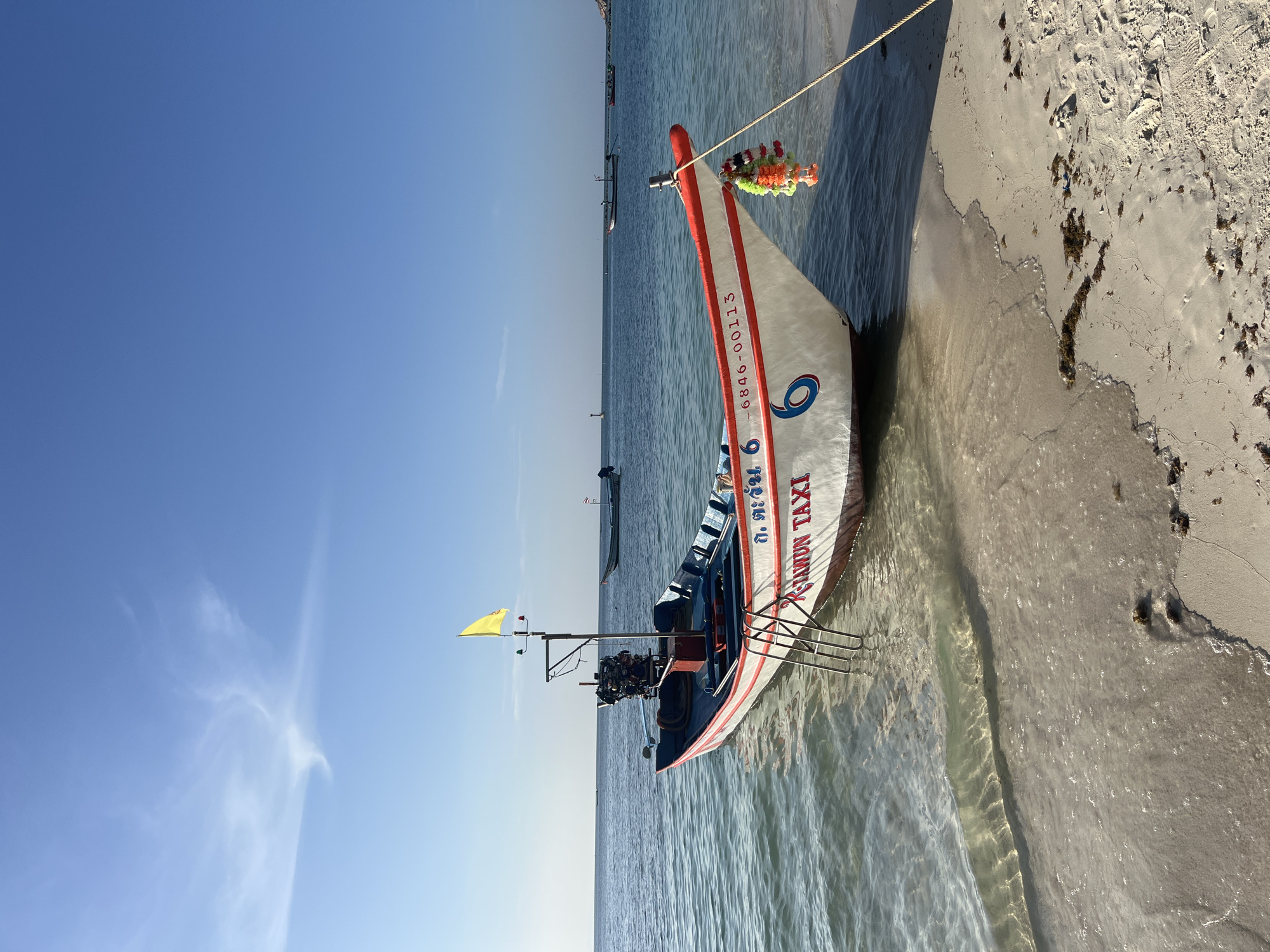 A longtail boat, identified as "TAWUN TAXI" and bearing the number "6", rests partially on a sandy beach and partially in the clear, shallow waters of Ko Pha-ngan District, Thailand. The boat is painted white with red trim and features a prominent outboard motor. A yellow flag flies from a mast on the boat, and decorative colorful garlands hang from its bow. The water exhibits a gentle ripple, and sunlight casts dappled patterns on the seabed visible through the clear water. In the distance, other boats are anchored on the calm, blue sea under a clear, bright sky with wispy clouds. The sandy beach is light-colored and appears relatively clean, with a few scattered pieces of seaweed. The overall scene depicts a tranquil coastal environment on a sunny day.