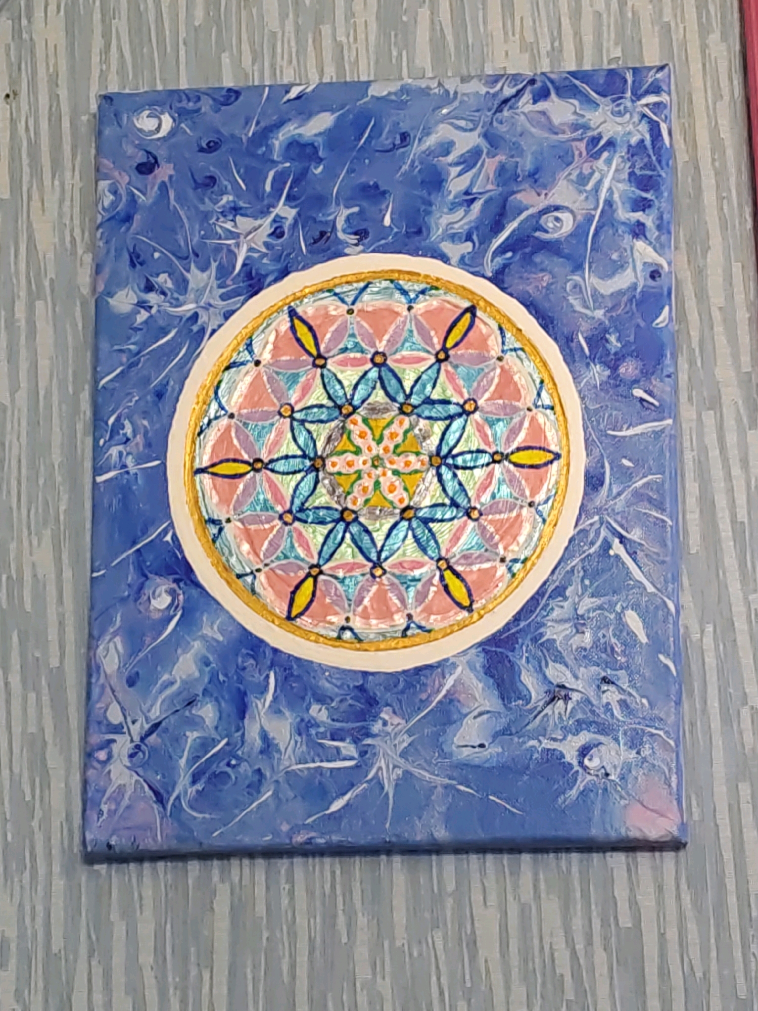 The image shows a painting with a floral geometric pattern against a blue, swirled background. The painting is on a canvas. The floral pattern is circular and enclosed within a gold ring. The flowers have petals in pink, blue, and yellow. The background is a marbled effect of blues and whites, with some hints of pink. The canvas is placed on a surface that appears to be a wooden tabletop or wall with a textured, light grey finish. The image is taken from a slightly elevated angle. There are no people or animals present in the image. The painting does not provide any specific clues about its location within Rostov-on-Don, Russia.