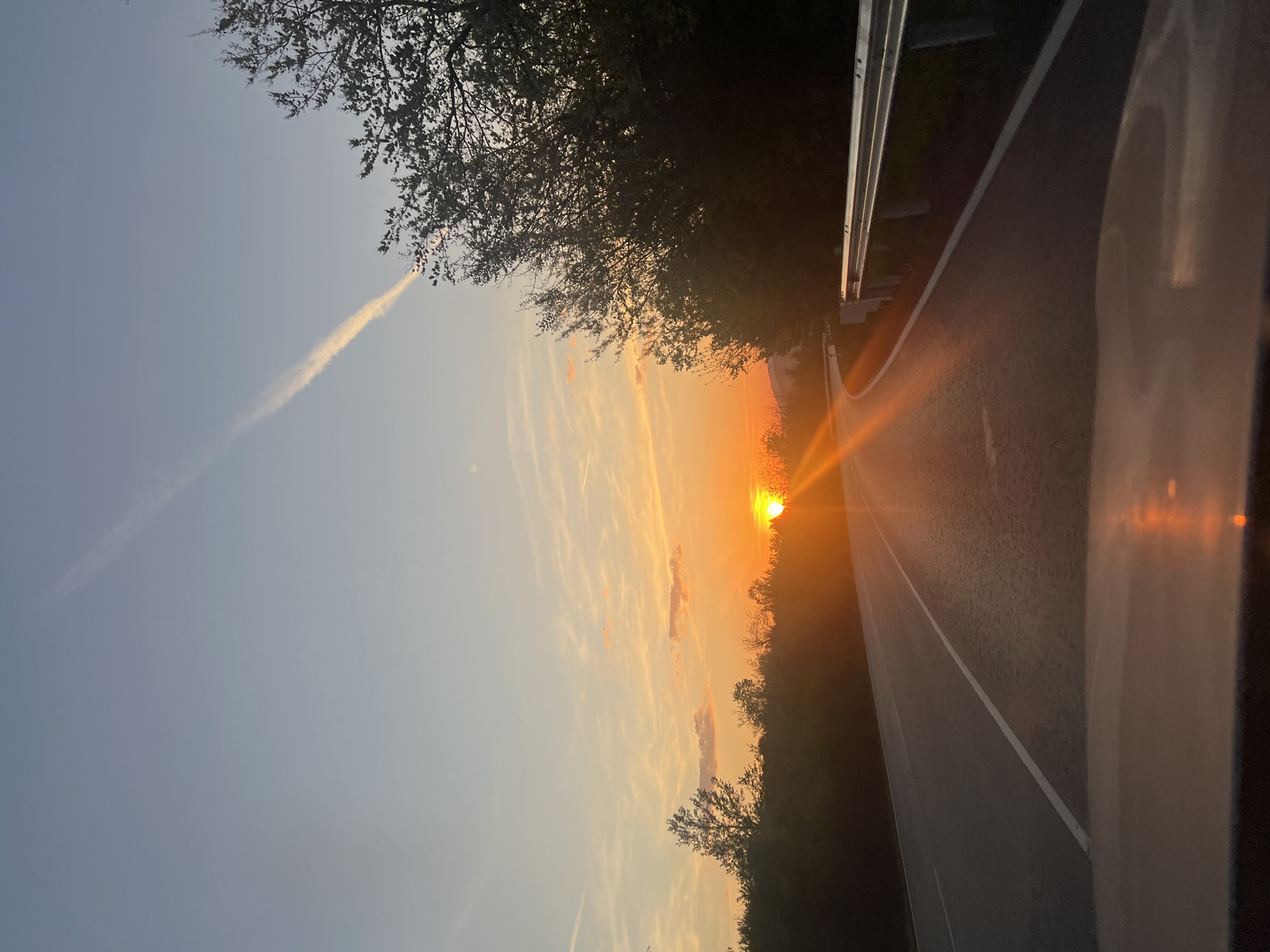 The image depicts a sunset over a road in Balkantsi, Bulgaria. The sun is setting directly ahead, creating a bright glare along the road's surface. A guardrail is visible along the right side of the road, bordered by trees. The sky is a mix of orange and blue hues, with contrails present. Part of a vehicle is visible in the foreground on the right side of the image.