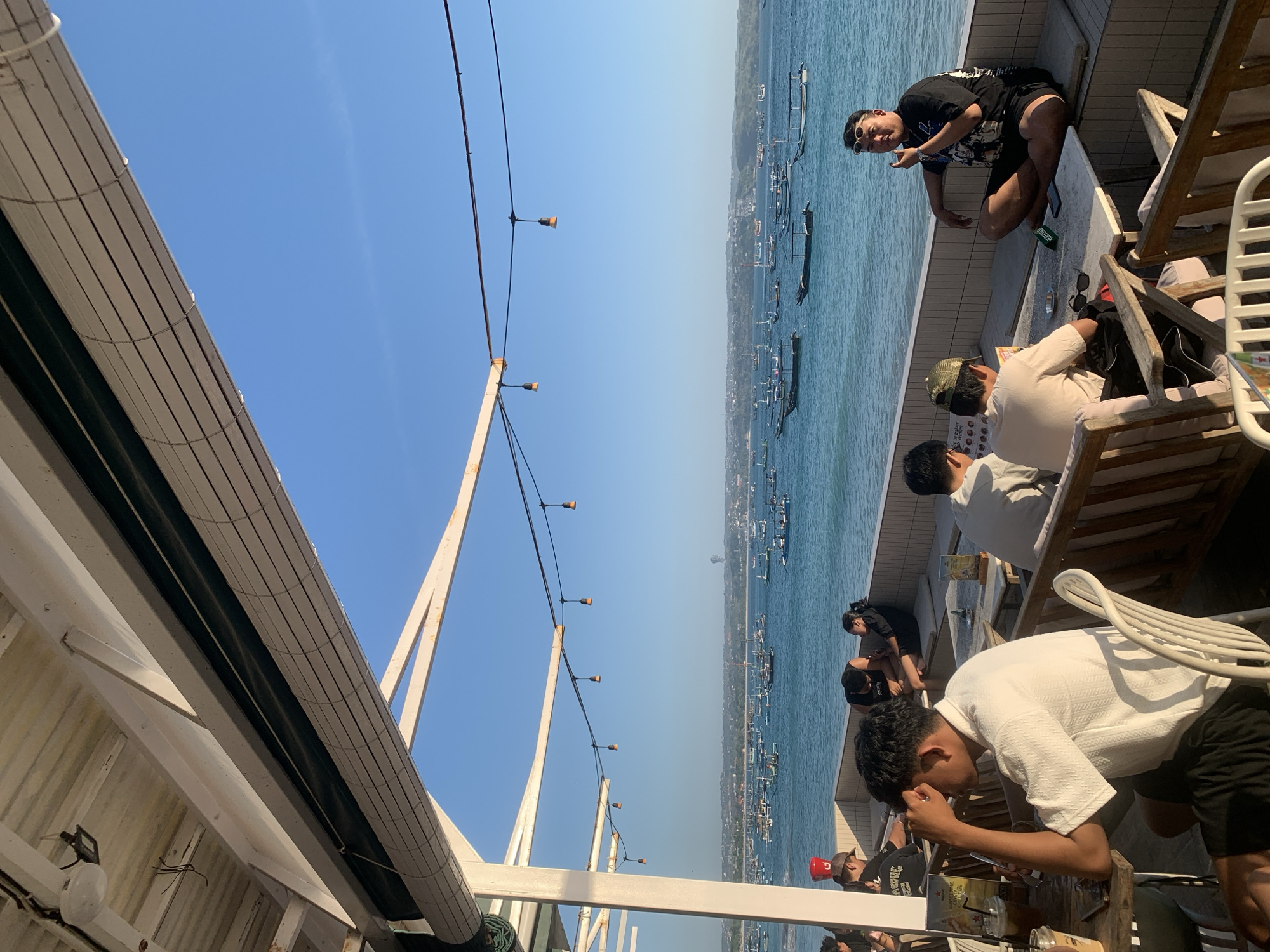 The image depicts a scene at a seaside establishment in Denpasar, Indonesia, likely a cafe or restaurant, overlooking a bay filled with numerous traditional Jukung boats. The establishment has an open-air structure with a white roof and support beams, decorated with string lights. Several individuals are seated at tables or on benches, enjoying the view. In the foreground, a man in a white shirt and black shorts is seated at a table, looking to his left. To his right, two other men are seated, one wearing a light-colored shirt and a green cap, and another in a white shirt. Further to the right, a man in a black t-shirt and black shorts, wearing sunglasses, is seated cross-legged on a bench, gesturing with his hand. The background shows a wide expanse of blue water dotted with many Jukung boats, and a distant coastline with buildings. The sky is clear and blue, suggesting it is daytime, likely late afternoon given the lighting.