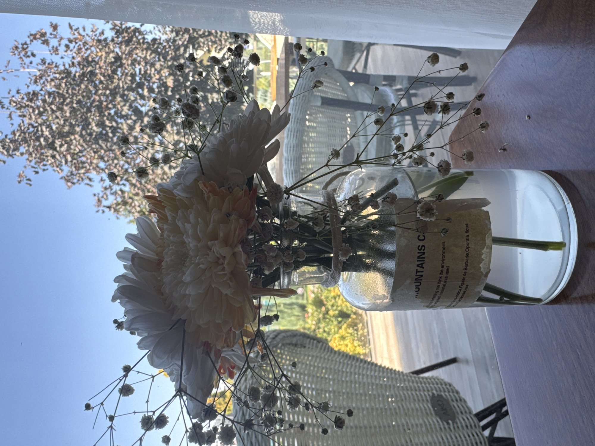 The image depicts a close-up view of a clear glass vase containing a bouquet of flowers and delicate baby's breath. The vase is placed on a wooden surface, likely a table, in what appears to be an outdoor or semi-outdoor setting. In the background, there are wicker chairs and tables visible, suggesting a café or restaurant patio. Beyond that, a large tree with autumnal foliage and a clear blue sky can be seen. A white sheer curtain is present on the right side of the frame, indicating proximity to an indoor space. A paper label is attached to the vase with some text that is partially legible, mentioning "MOUNTAINS C". The overall lighting suggests it is daytime, possibly late morning or afternoon, with sunlight filtering through the scene. There are no people visible in the image, and the focus is on the floral arrangement and its immediate surroundings. The scene does not provide any specific indicators for a location within Tashkent, Uzbekistan.