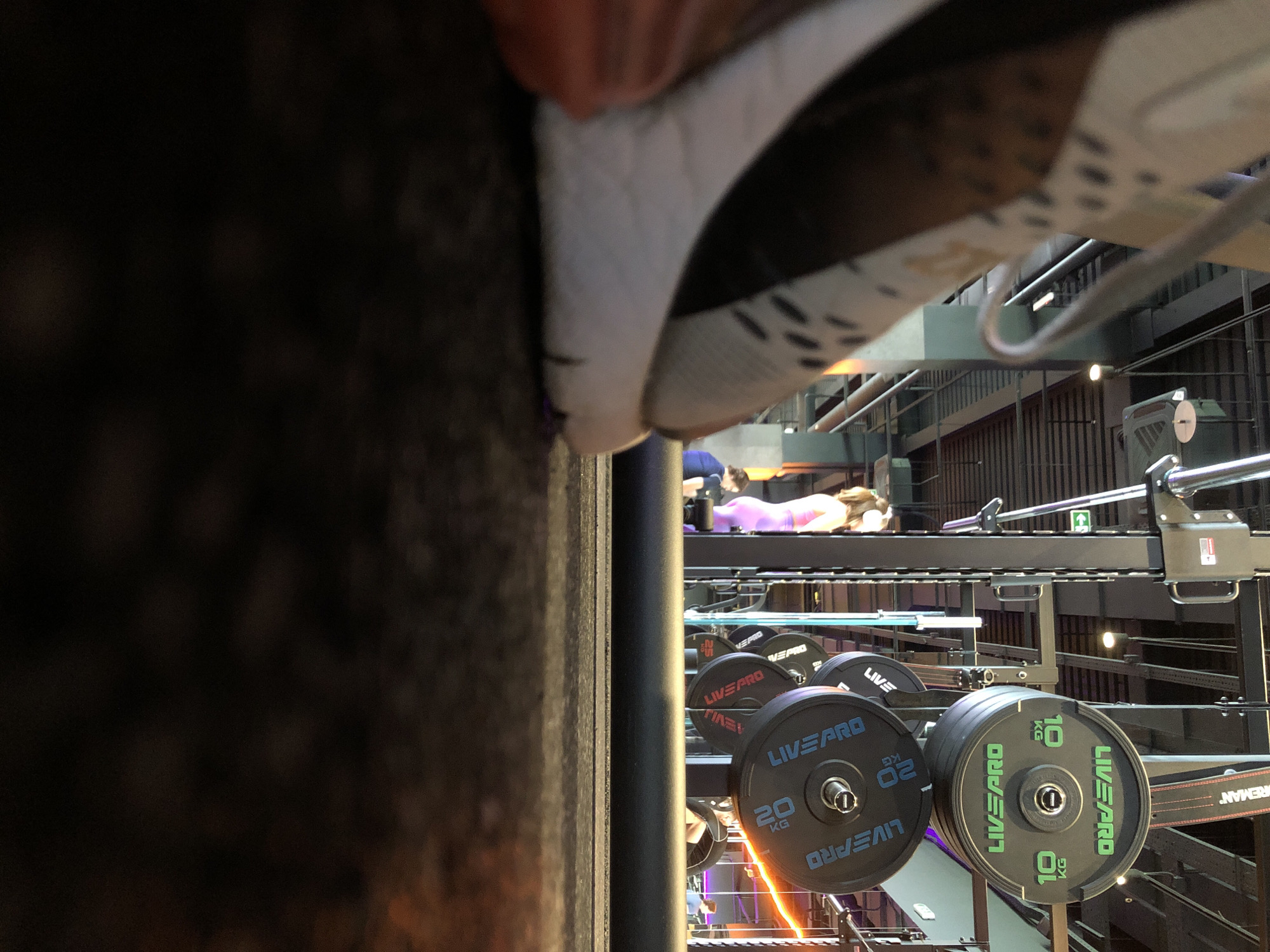 This image presents a slightly tilted, low-angle view inside a modern, multi-level gym or fitness center, located in Krasnoyarsk, Russia. The immediate foreground is occupied by a blurred close-up of a shoe, featuring a white sole and a dark upper, obscuring much of the left side of the frame. The perspective suggests the photo might have been taken from a first-person point of view or with the camera placed on a surface next to a shoe.
In the midground and background, various gym equipment is visible, primarily black barbell weight plates from the brand "LIVEPRO." Several plates clearly show their weight denominations, including "20 KG" and "10 KG." These plates are racked on metal structures, which also include bars and other gym apparatus. The gym's architecture features dark metal railings and supports, indicating a multi-story layout. In the upper background, a person can be seen lying on a piece of equipment, possibly engaged in an exercise or resting. Partially visible text "REMAN" can also be observed on some equipment. The setting is well-lit by interior lighting, suggesting an active gym environment.