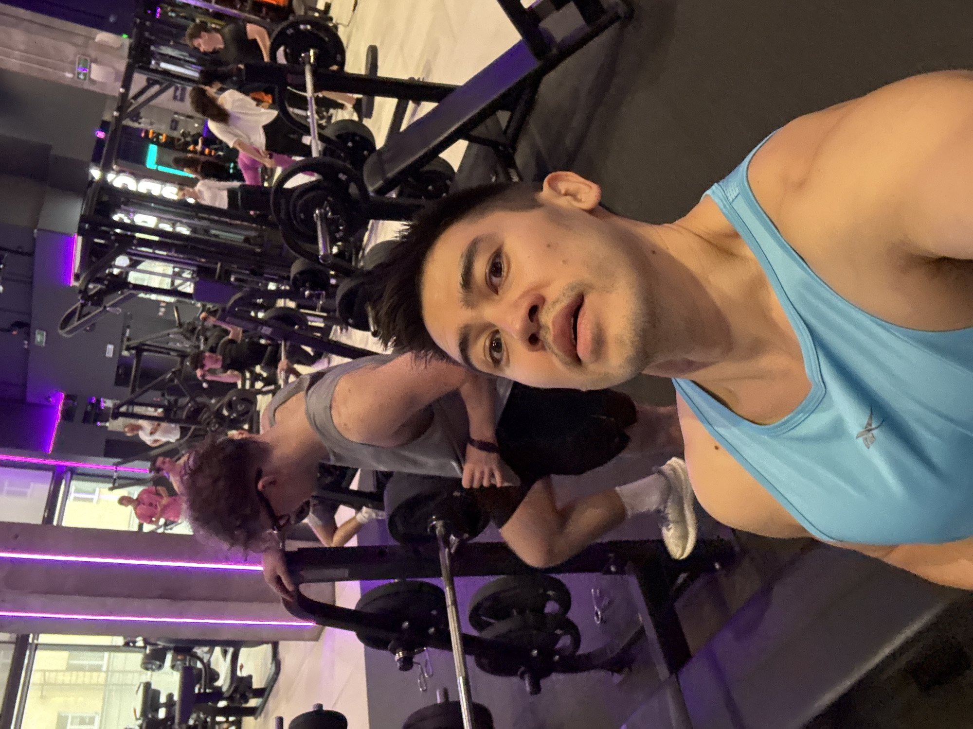 This image depicts an interior scene from a modern gym in Kazan, Russia. The primary subject is a man in his late 20s or early 30s, positioned in the foreground, taking a selfie. He has short dark hair, a light mustache, and is wearing a light blue athletic tank top, looking directly into the camera with a neutral to slightly pensive expression.
The background shows a busy gym floor with several individuals engaged in workouts. Various pieces of gym equipment are visible, including multiple squat racks, barbells with weight plates, and benches. Closer to the foreground, a man with curly dark hair and glasses, dressed in a grey t-shirt and dark shorts, is leaning on a piece of equipment. Further back, another person in a white t-shirt and pink leggings is visible near a squat rack.
The gym's ambiance is characterized by contemporary design, featuring prominent purple LED strip lighting that adds a vibrant glow to the space. Large windows in the background offer a view of an urban setting with other buildings, suggesting the gym is located in a city area. The scene captures typical activity during a workout session. No distinct text is clearly visible in the image.