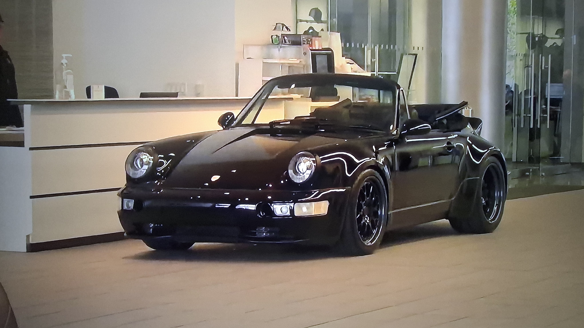 The image captures a black Porsche 911 convertible parked on a light-colored floor that resembles a showroom or dealership. The car's sleek design is highlighted by its wide fender flares. In the background, there's a reception desk and a shelving unit displaying various items. Large glass doors and a supporting column are visible, suggesting a modern building in an urban environment. A person is partially visible on the left side of the frame. The location in the city of Unknown, Unknown is indeterminate given the available information.