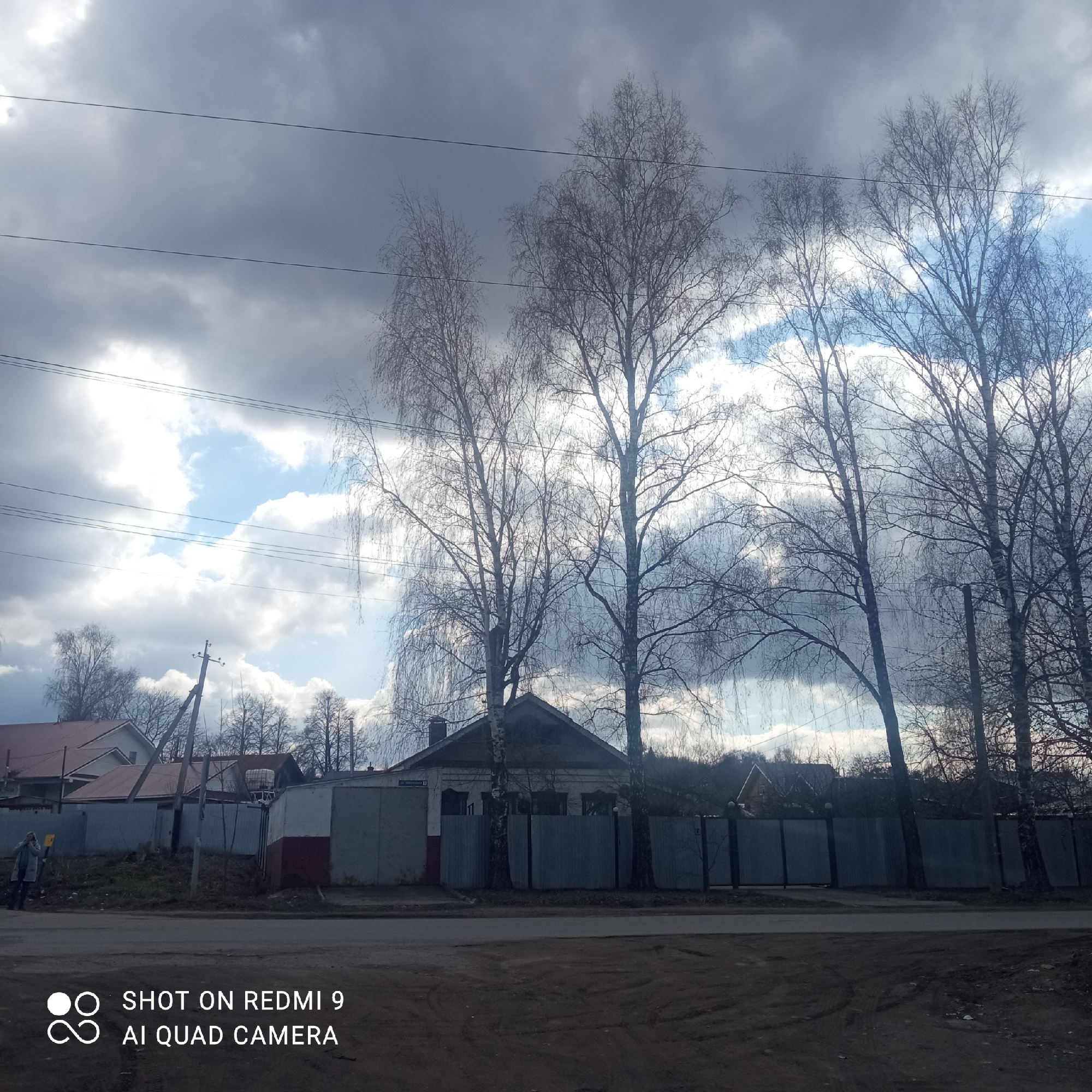 The image depicts a street scene in Chaikovsky, Russia. The sky is overcast with a mix of dark gray and white clouds, and there are leafless birch trees lining the street. In the background, there are houses with fences and some structures that appear to be sheds or garages. In the foreground, there is a paved street and a dirt shoulder. A person is walking on the sidewalk in the distance on the left side of the image. An electrical power pole can be seen in the background. The photo was taken with a Redmi 9 AI Quad Camera.