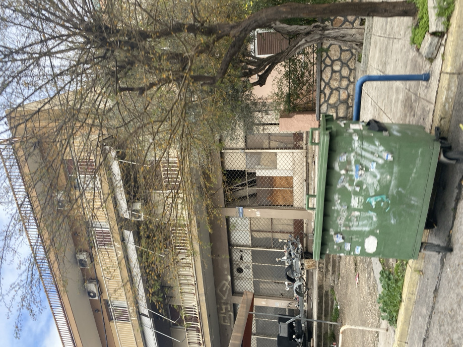 This image captures a static, daytime urban street scene in Nikea, Greece, characterized by an aging multi-story building, a large tree, and a prominent green dumpster.
The main subject is a concrete building, likely residential or mixed-use, with at least three visible floors. It features numerous balconies with white metal railings, and several outdoor air conditioning units are mounted below the windows on the upper floors. The ground floor of the building appears to be a disused commercial space or entrance, with large window and door openings covered by dark metal chain-link fencing or security grates. Some of these ground-floor openings are boarded up with plywood, and there is dark, illegible graffiti spray-painted on the facade.
In front of and partially obscuring the building, a large, deciduous tree dominates the upper and central parts of the frame. Its branches are mostly bare with only sparse, light-colored leaves, suggesting either late autumn, winter, or early spring.
To the right, positioned on a concrete sidewalk, is a large, wheeled green dumpster. Its surface is heavily covered with a mix of white and blue graffiti, stickers, and torn paper, making any specific text unreadable. A bright blue pipe emerges from the ground near the dumpster and bends upwards.
Behind the tree and the dumpster, a stone wall with a distinctive textured, possibly decorative, pattern is visible, alongside what appears to be another building or part of a structure with a window fitted with dark wooden shutters. The sidewalk consists of grey concrete slabs, with some weeds growing in cracks. The overall impression is that of an older, lived-in urban environment, perhaps somewhat neglected. No people or activity are visible. The lighting suggests it is daytime, possibly overcast.