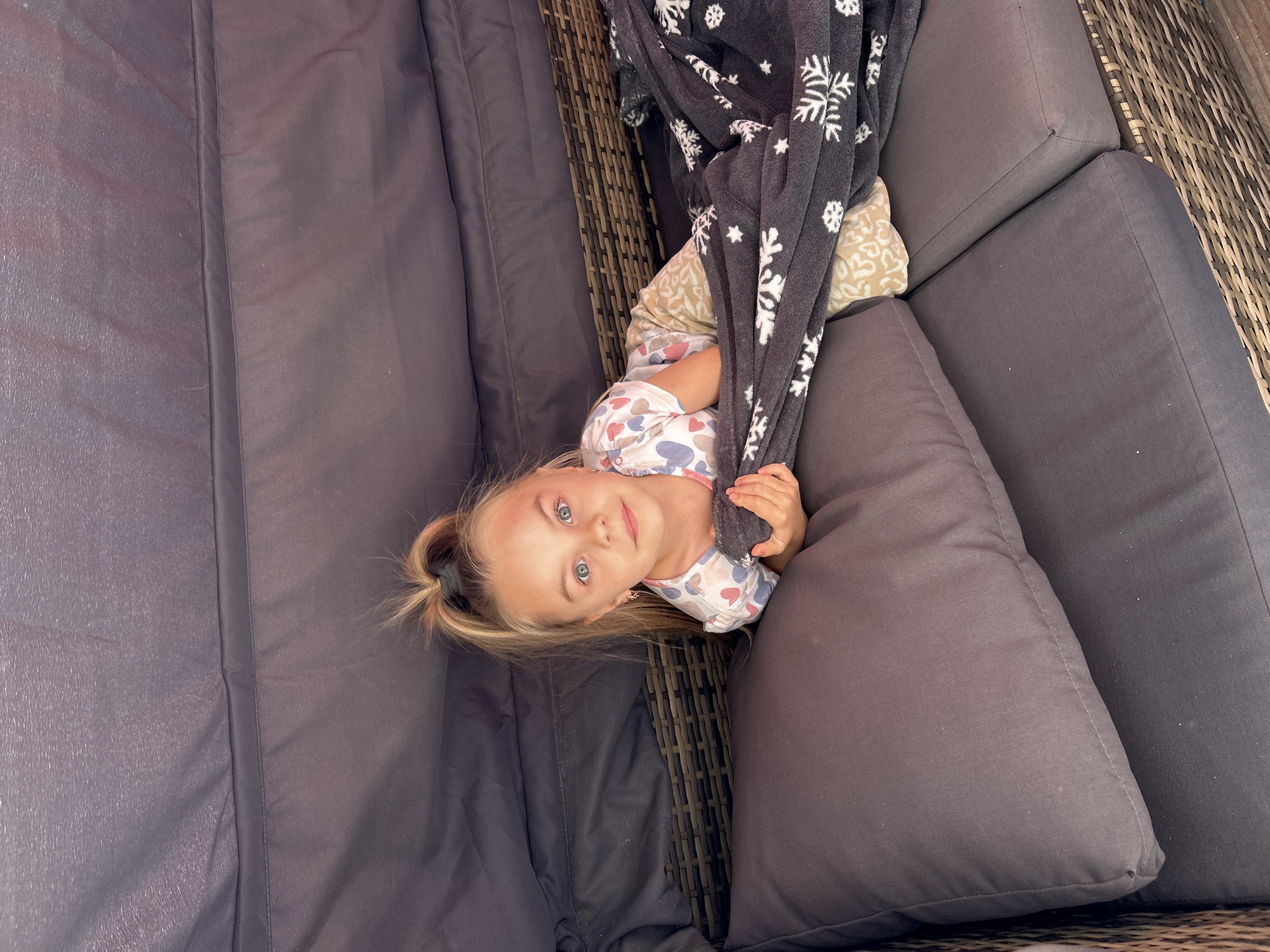 The image shows a young girl lying on a dark-colored outdoor sectional sofa. She is partially covered by a dark gray blanket with white snowflake patterns. The girl has blonde hair tied in a ponytail and is wearing a light-colored pajama set with heart patterns. She is looking directly at the camera with a gentle expression. The sofa is upholstered in a dark fabric and has thick cushions. The background shows a wicker weave pattern, likely part of the sofa's frame or surrounding furniture. The lighting suggests it is daytime, possibly outdoors given the furniture style. The girl is holding onto the blanket, seemingly relaxed or playing. There is no visible text in the image. The location is specified as Waldbröl, Germany.